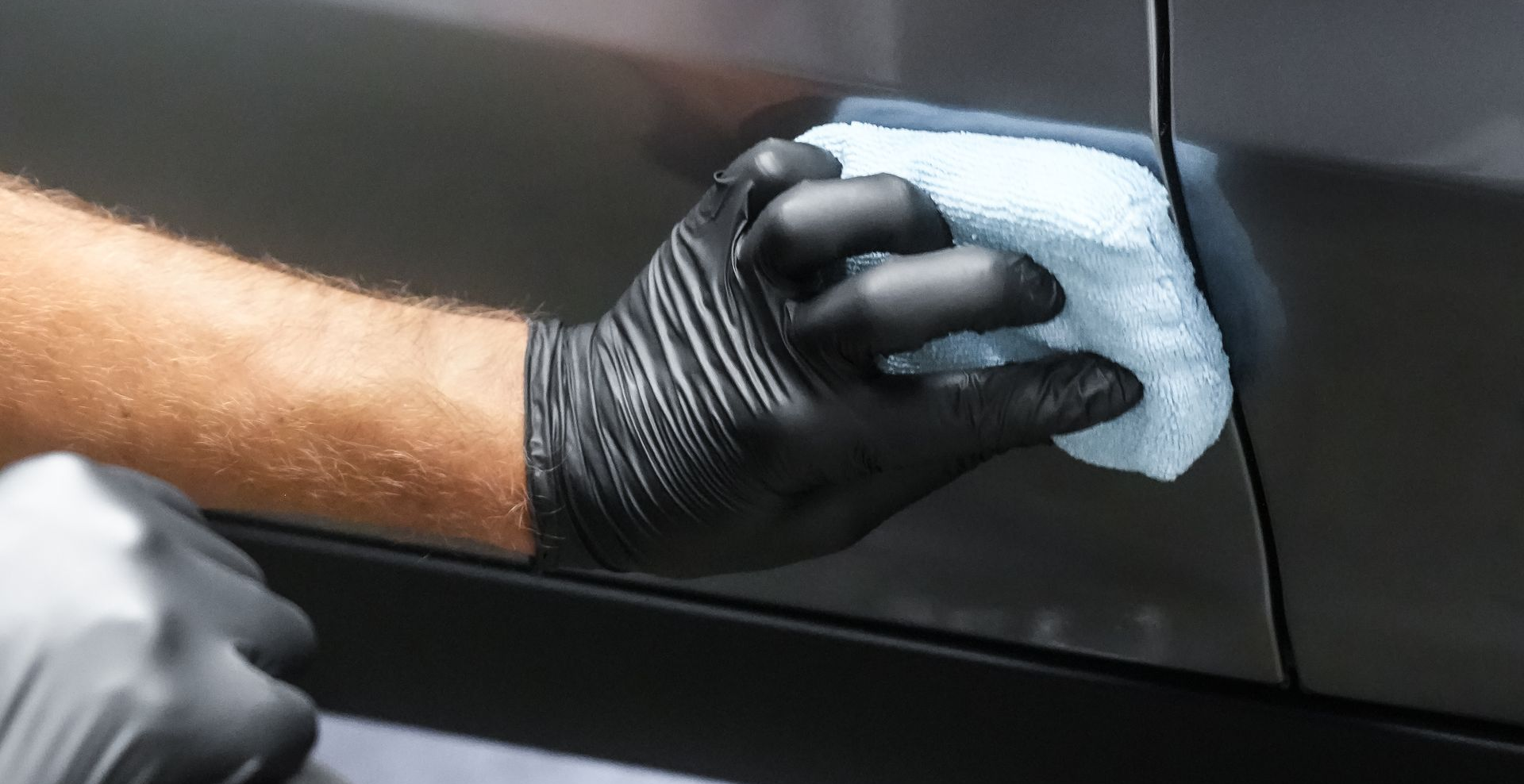 Gloved hand wiping a car surface with a microfiber cloth.