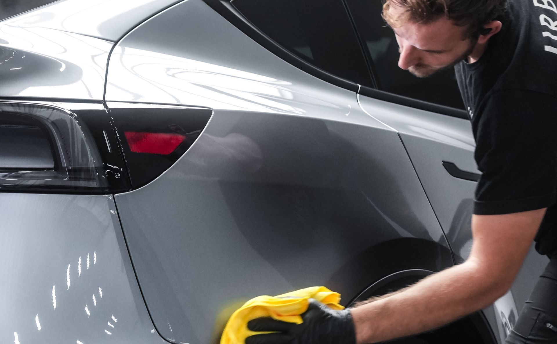 A person wearing gloves wipes a gray car with a yellow cloth.