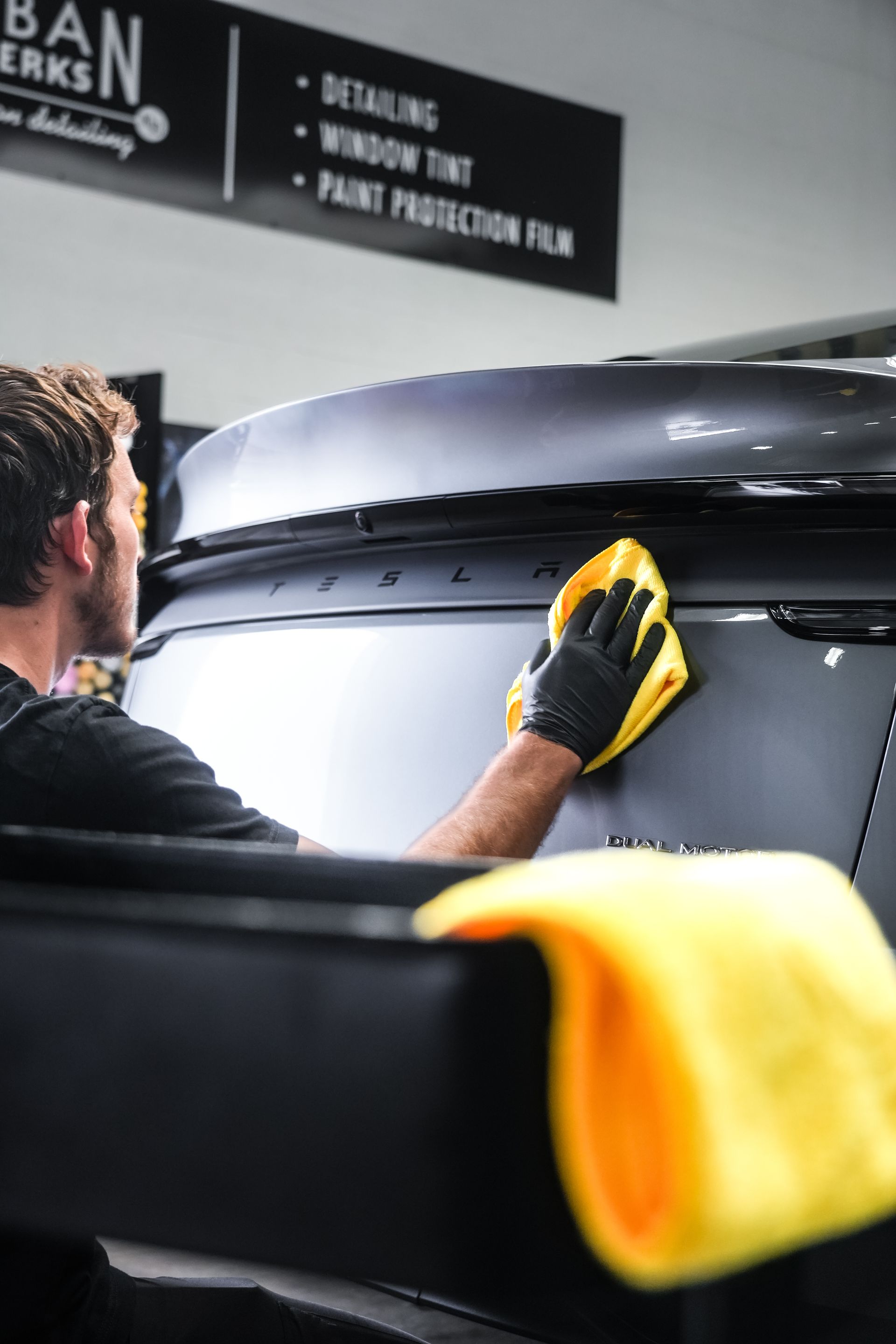 Man wearing black gloves, cleaning a dark vehicle with a yellow cloth. Sign lists detailing services.