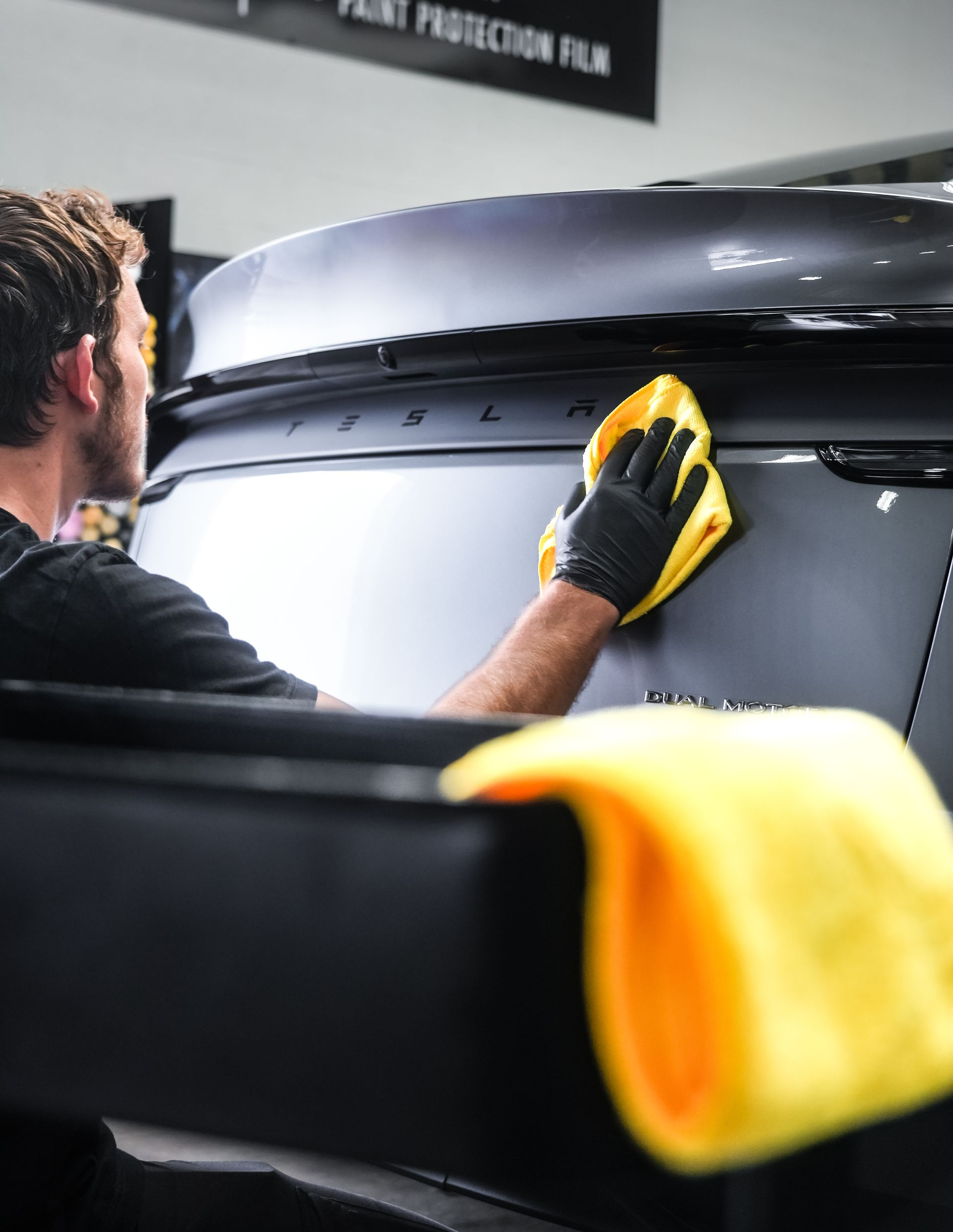 Person in black gloves wiping a dark car with a yellow cloth.