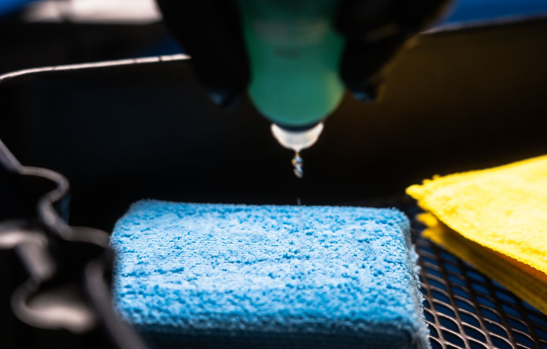 Blue sponge being squeezed with liquid from a bottle, next to yellow cloth.