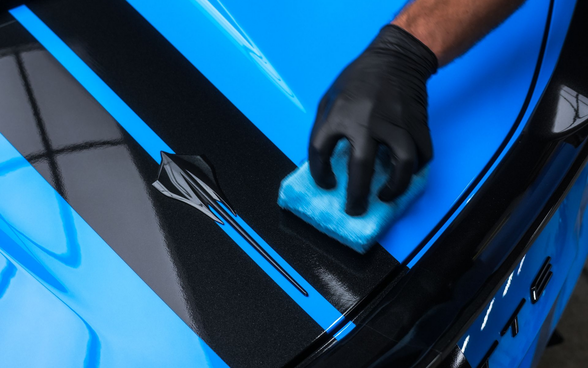 Gloved hand using blue sponge to clean a blue and black car hood with racing stripes.