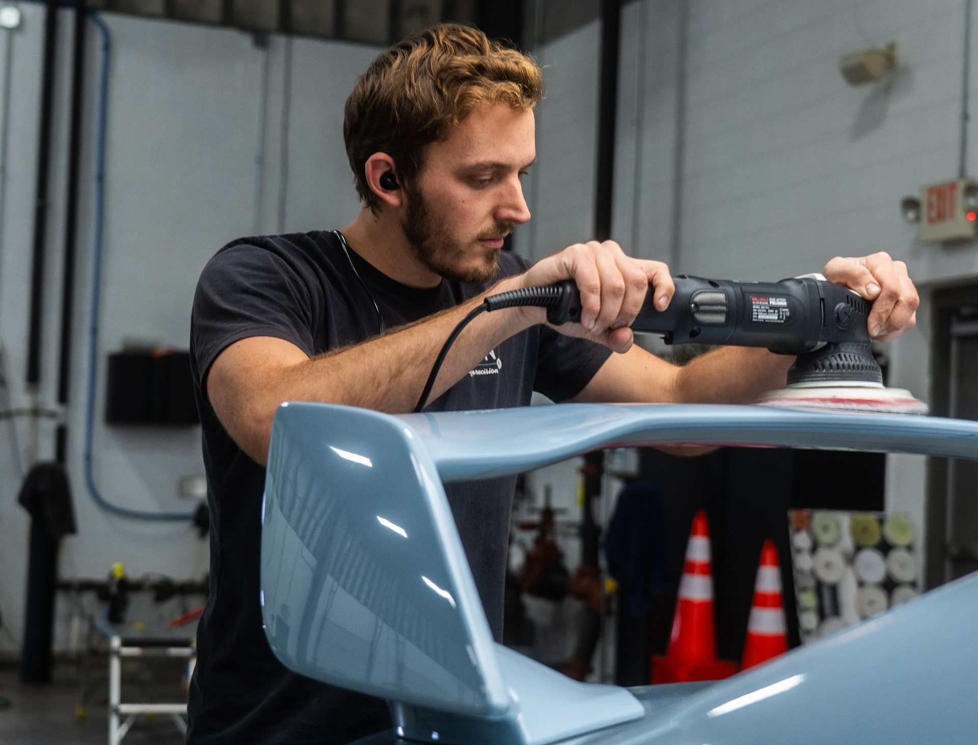 Man polishes a light blue car spoiler with an electric buffer in a workshop.