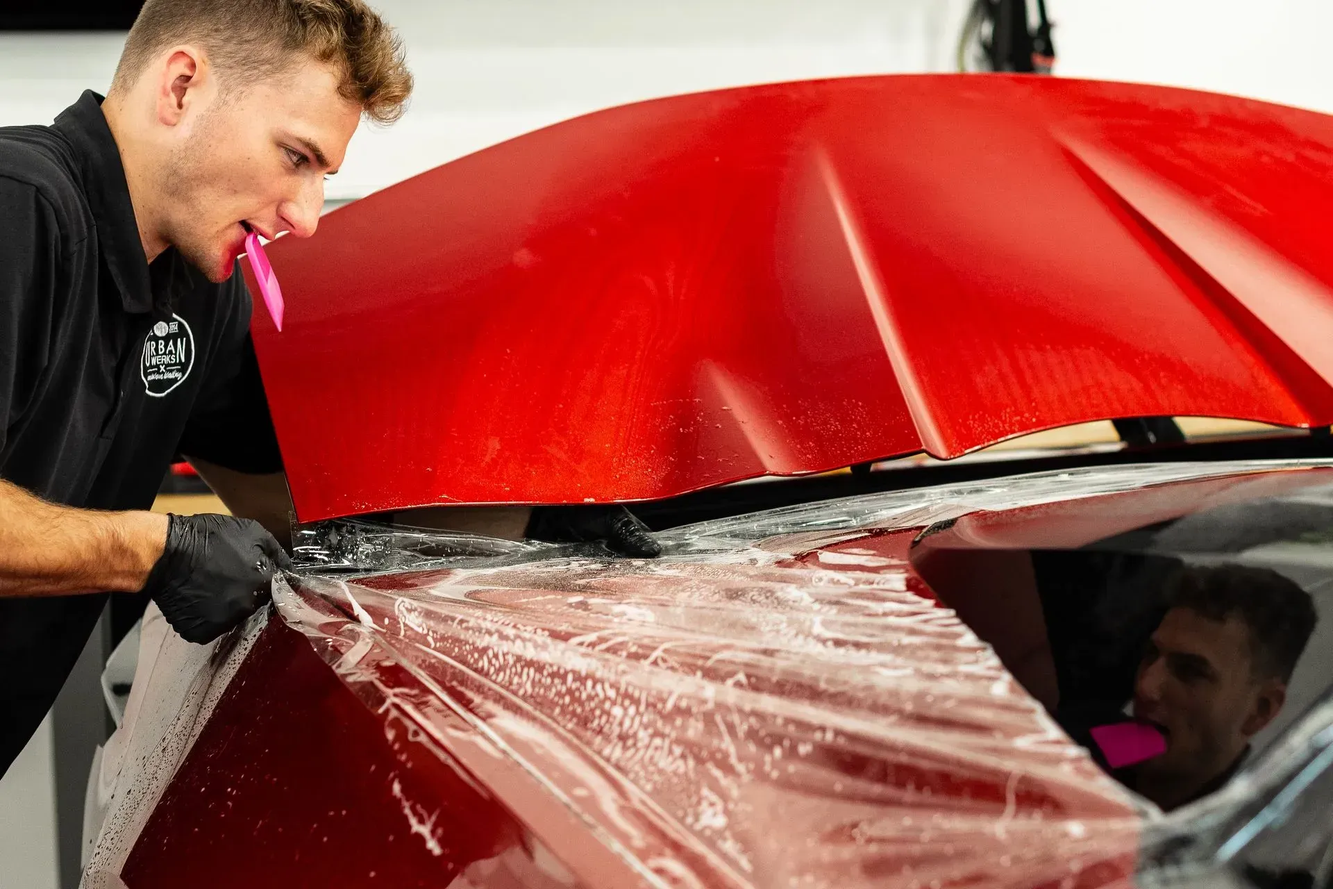 Man using a pink tool to install protective film on a red car hood.