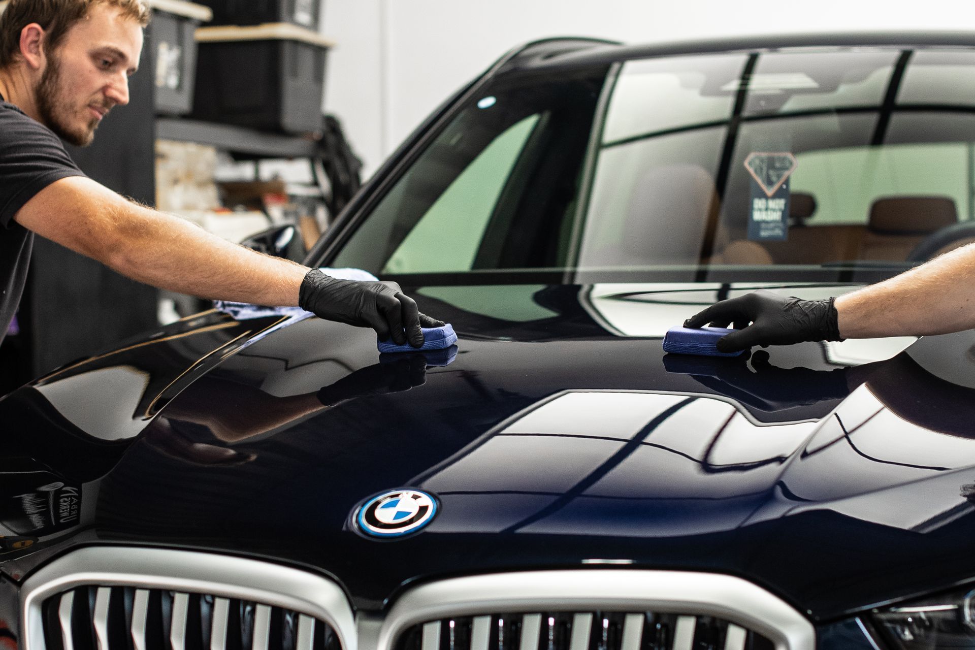 Two people applying ceramic coating to a navy blue BMW hood in a garage.