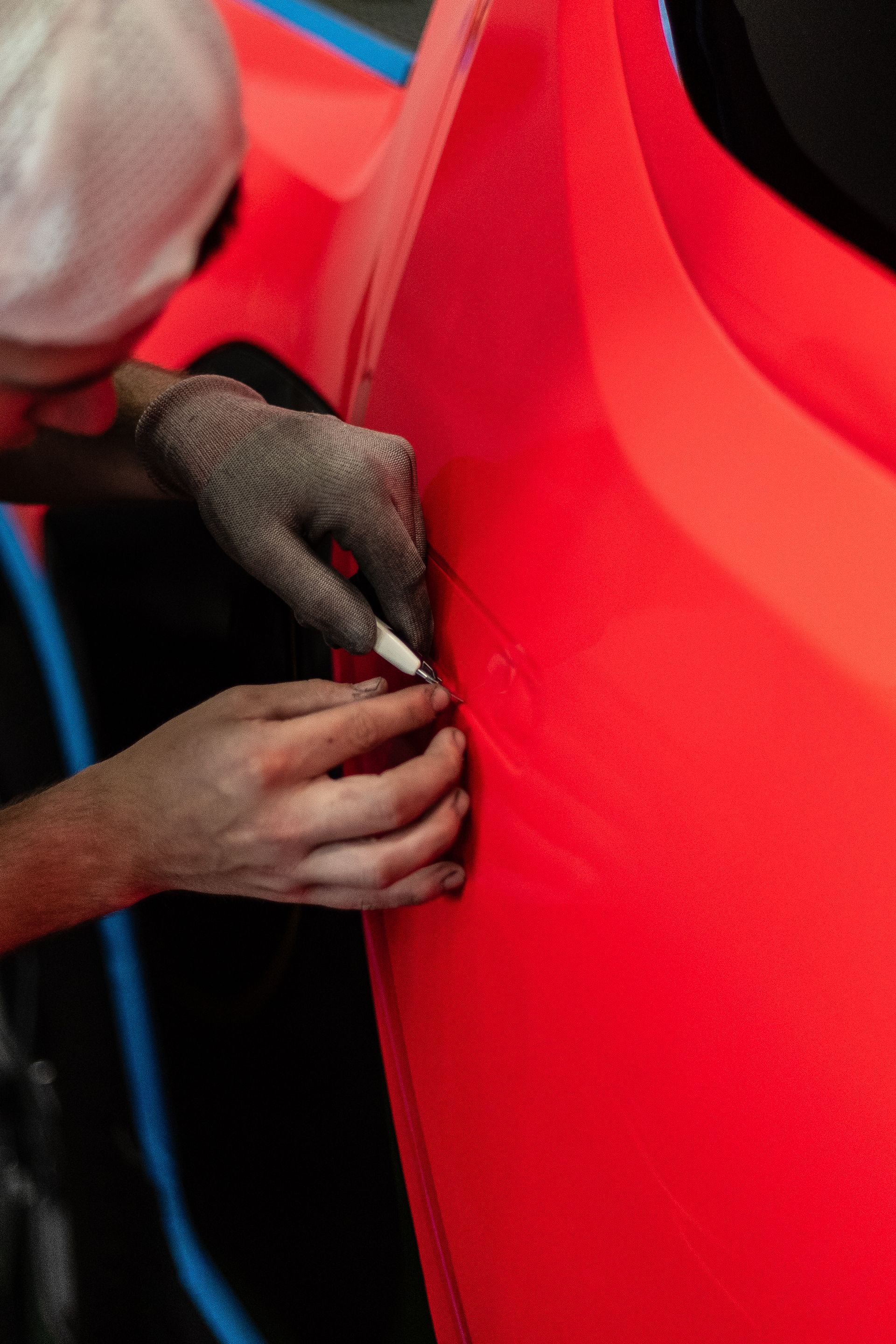 Person using a tool on a bright red car panel, wearing a face mask and dark gloves.