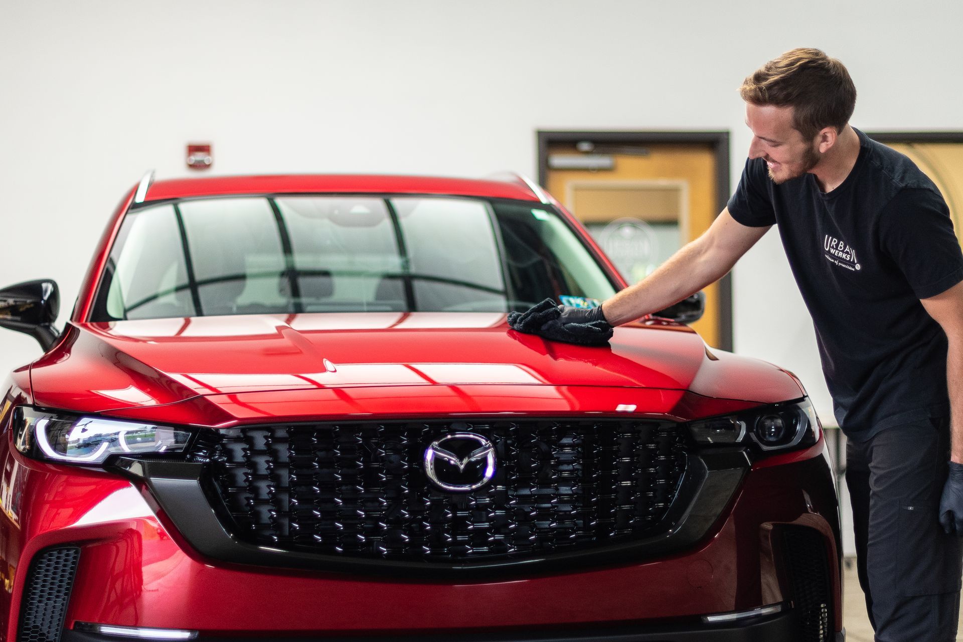 Man cleaning a shiny red Mazda SUV's hood in a garage.