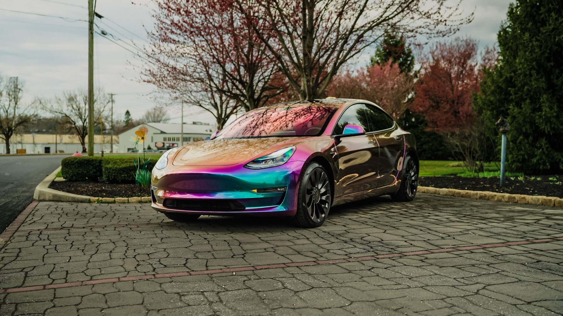 Tesla car with iridescent wrap parked on cobblestones. Cherry blossoms in the background.
