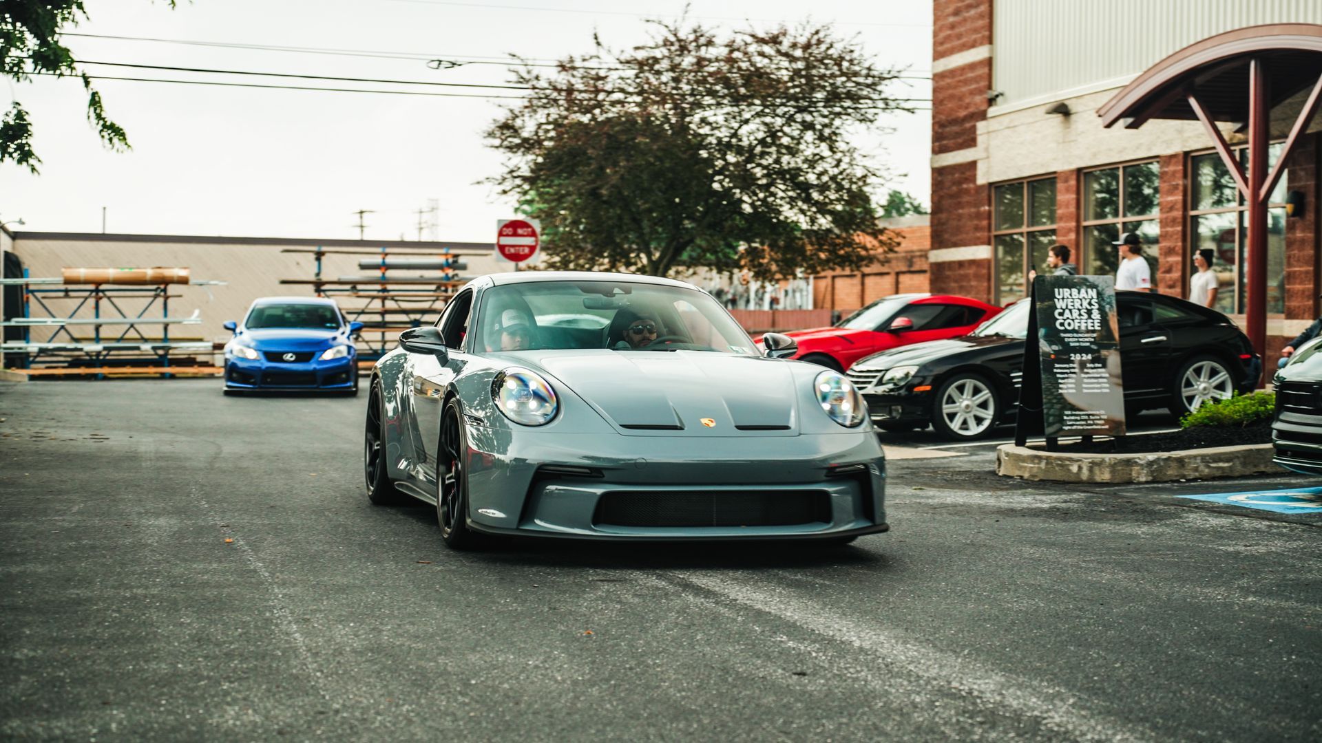 Gray Porsche sports car driving on a paved lot, other cars and a building in the background.