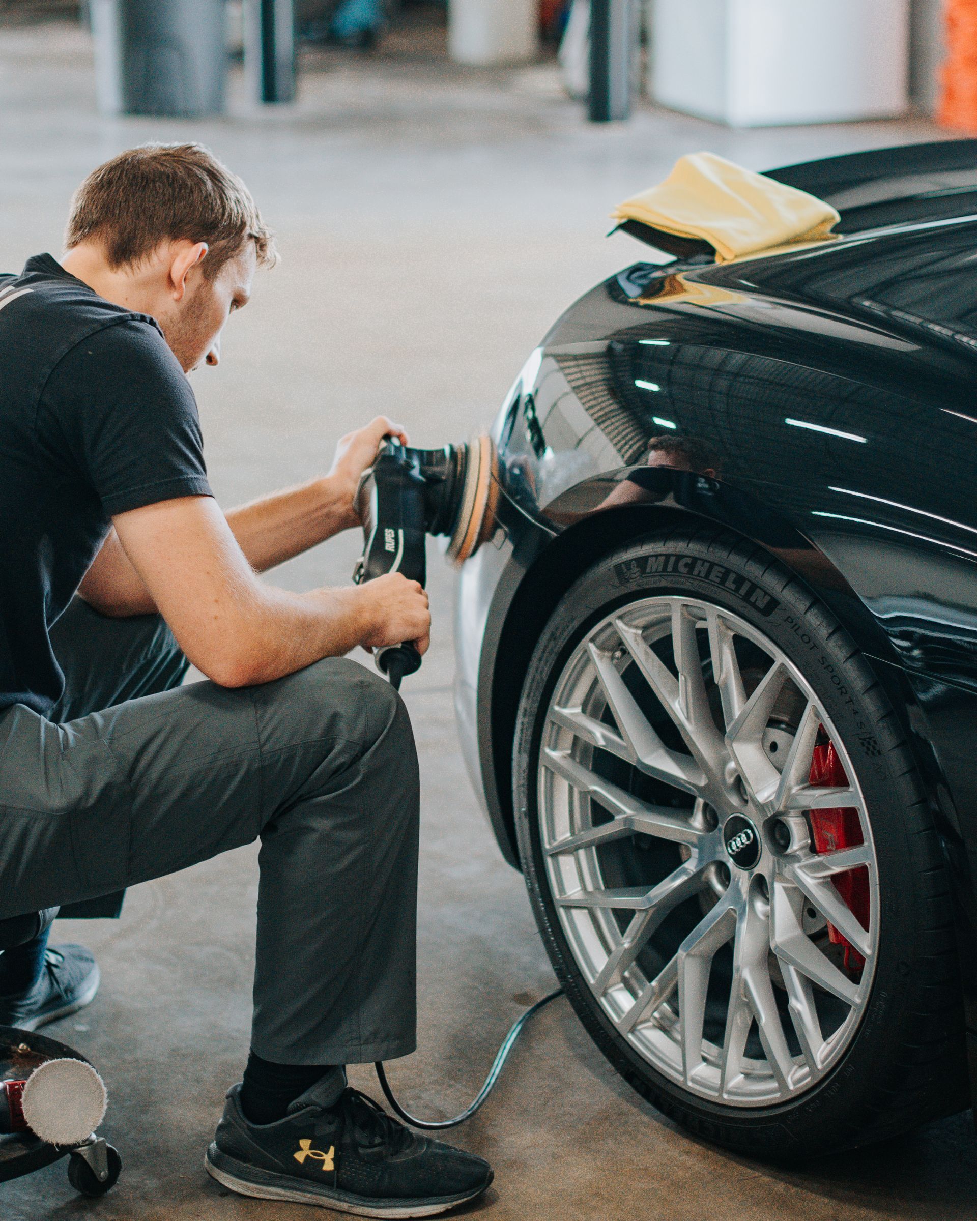 Man polishing a black car's front fender with a power buffer. Shiny wheel and red brake caliper visible.