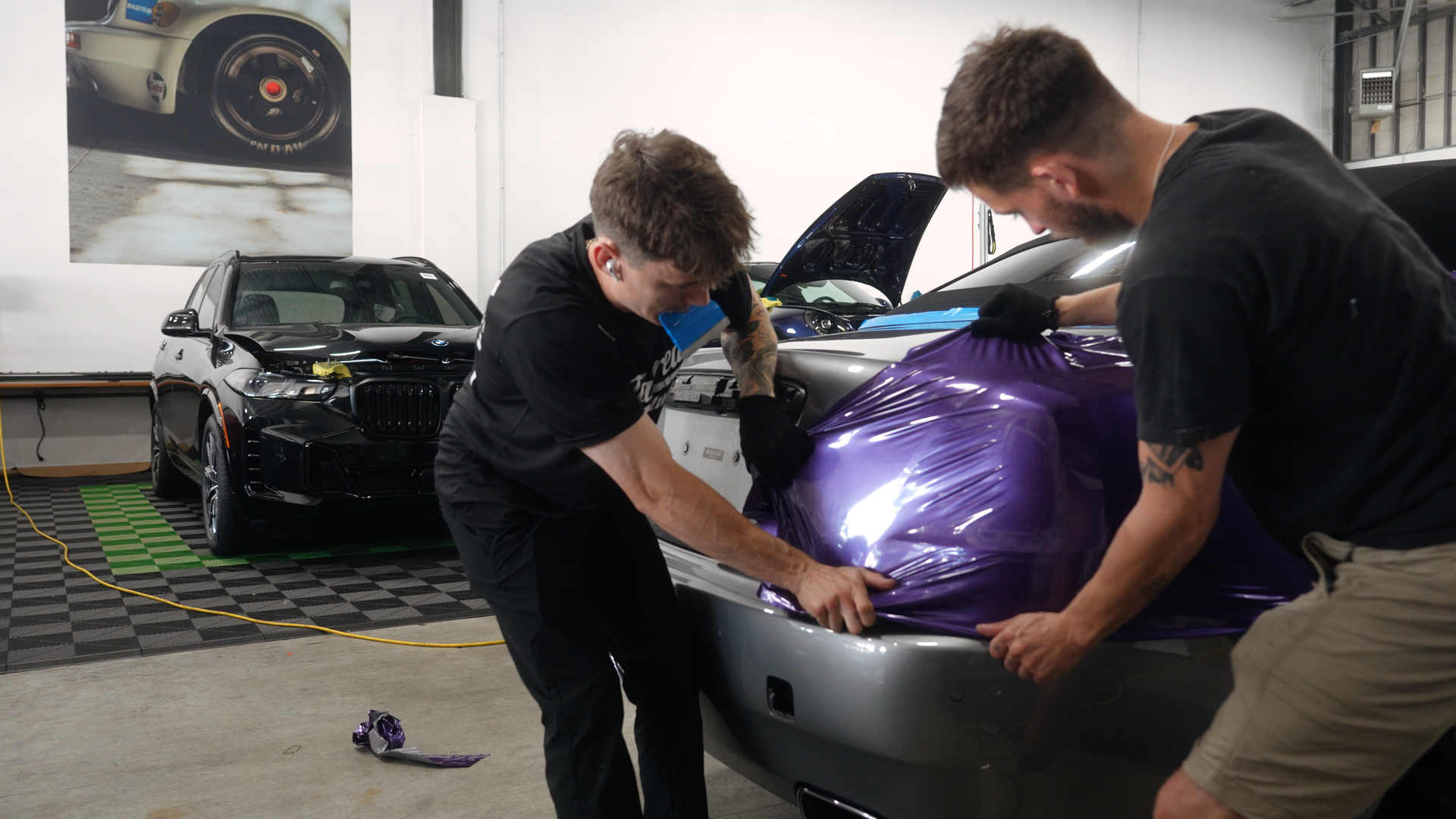 Two people wrap a car bumper in purple vinyl at a shop.