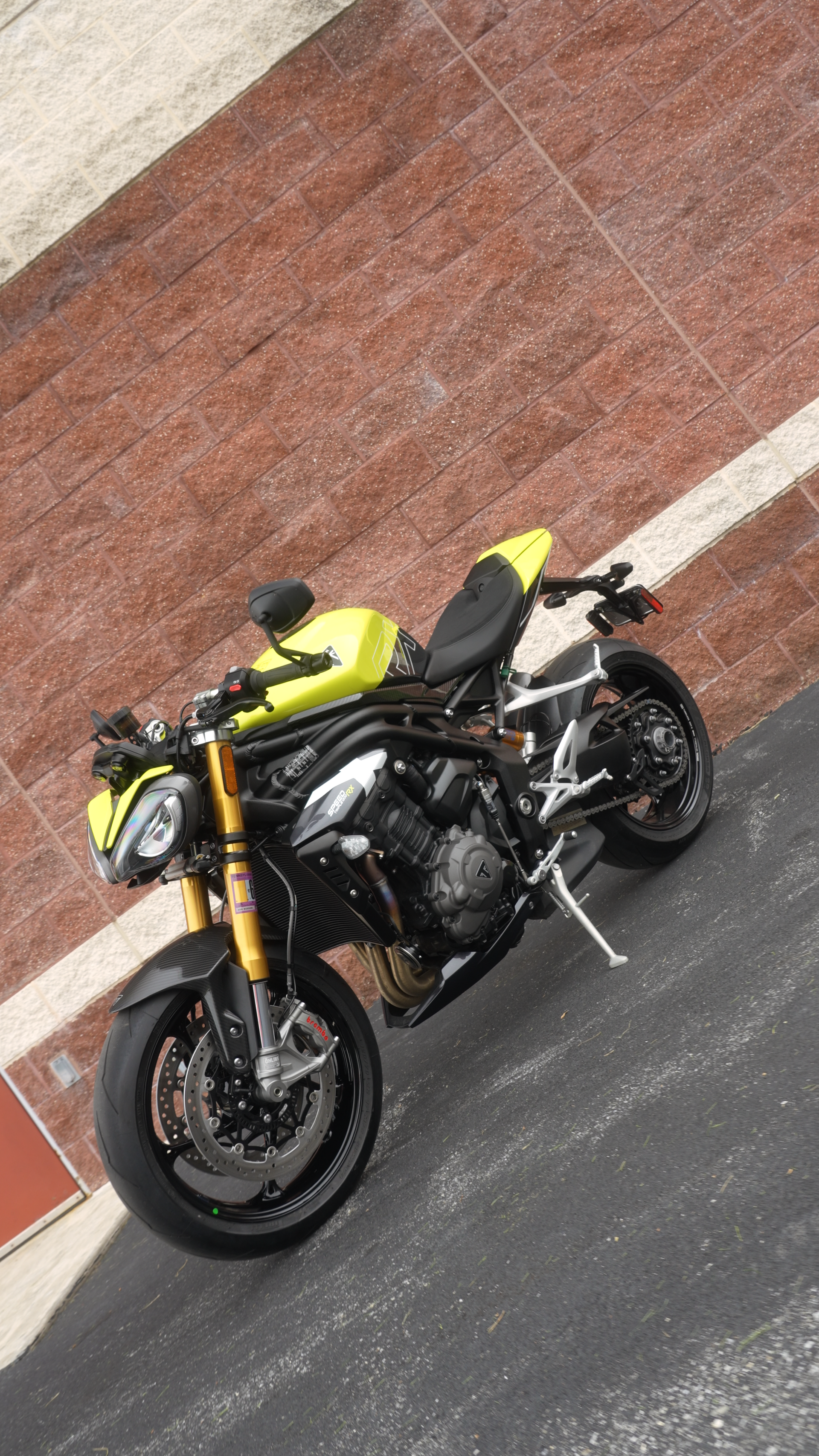 Yellow and black Triumph Speed Triple motorcycle parked on asphalt in front of a brick wall.