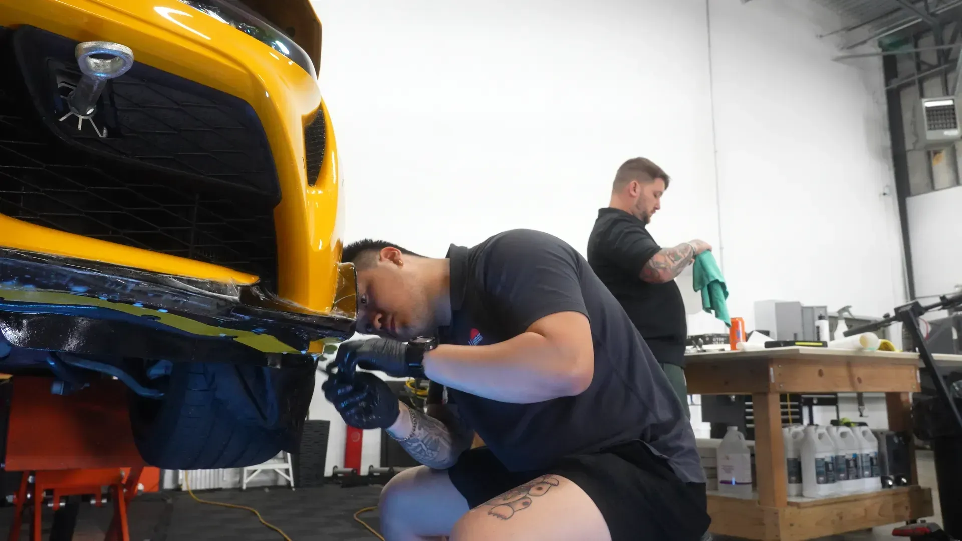 Two mechanics working on a yellow car in a garage. One is kneeling, focused on the front bumper.