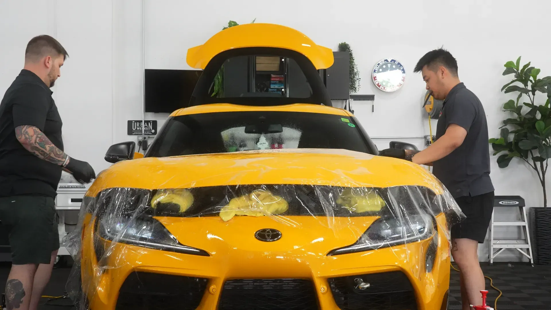 Two people applying protective film to the front of a yellow sports car in a garage.