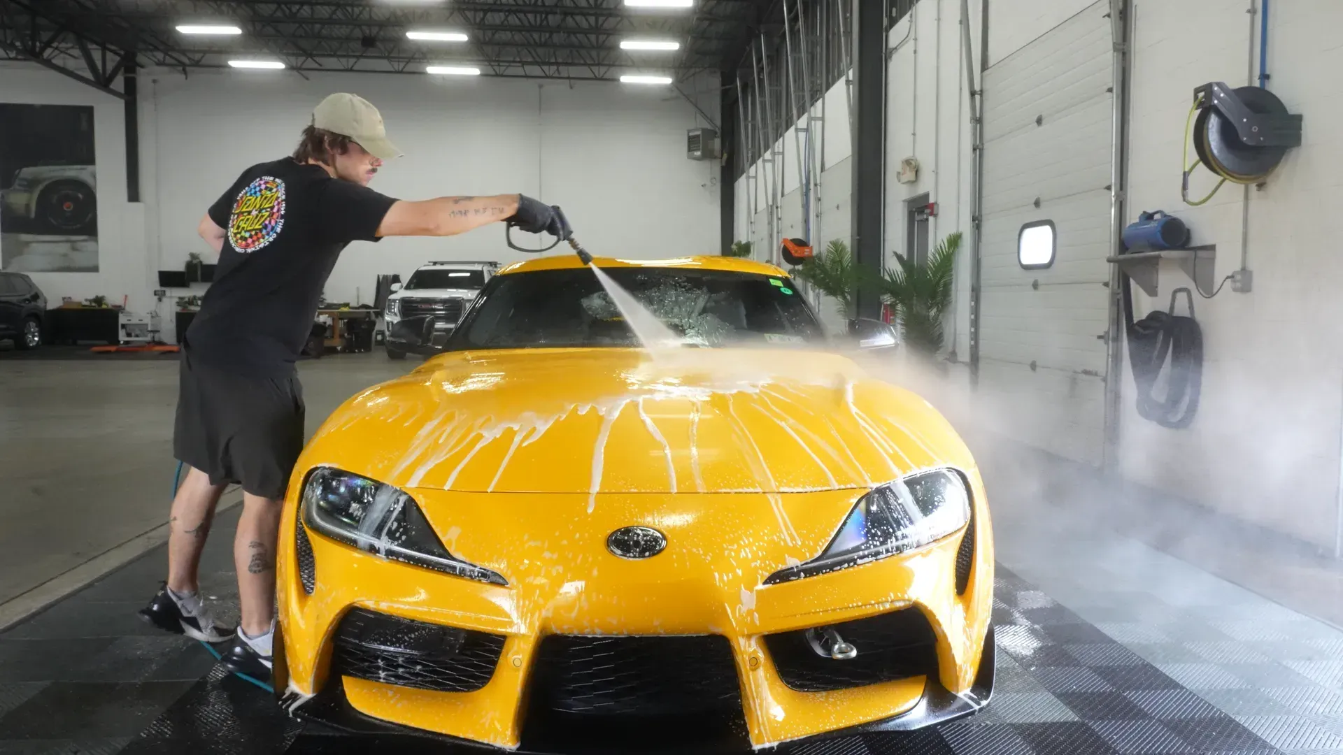 Man washing a bright yellow sports car with a pressure washer inside a garage.