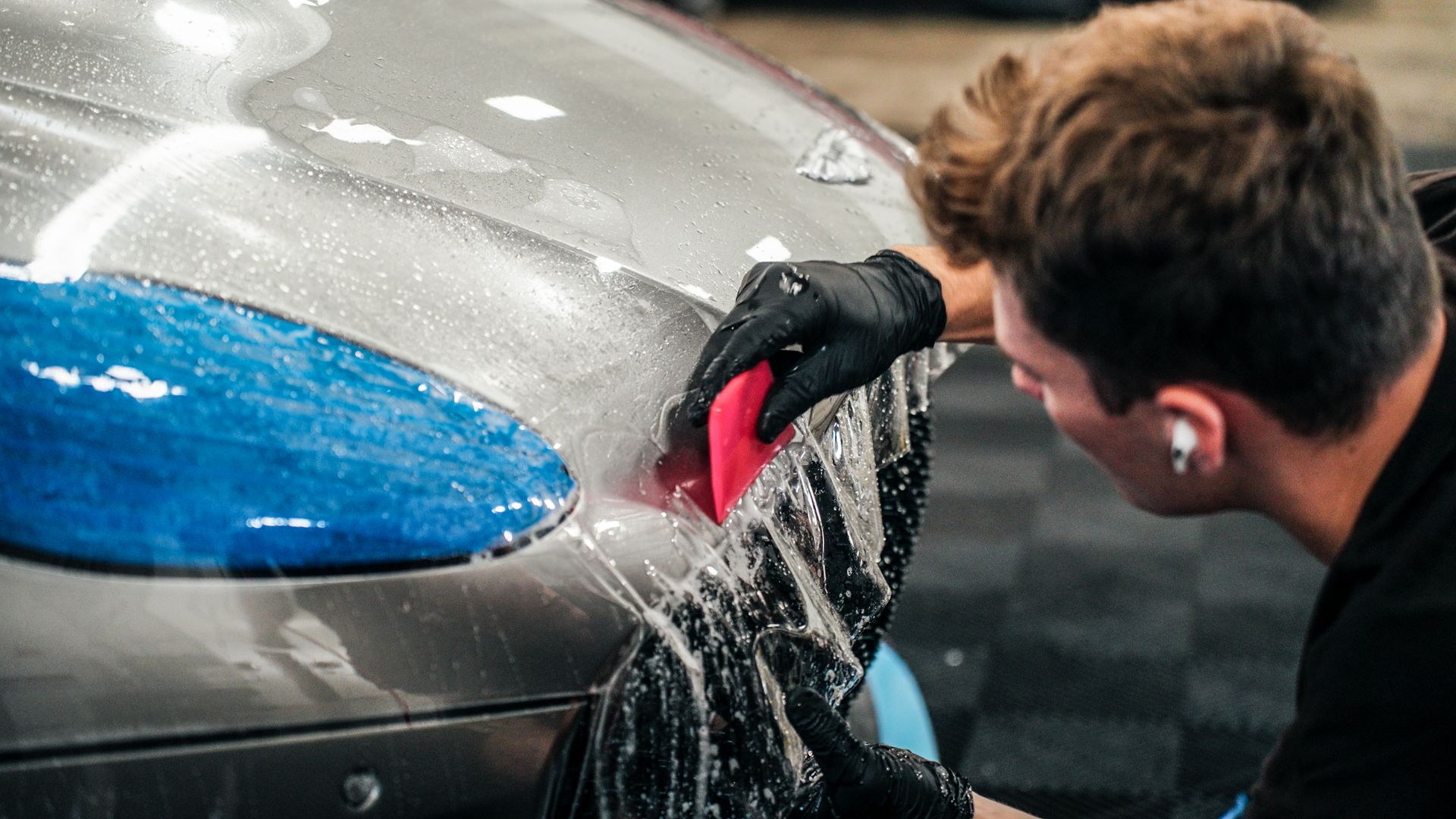 Man in gloves applying film to a car's hood, using a squeegee. Wet, silver car.
