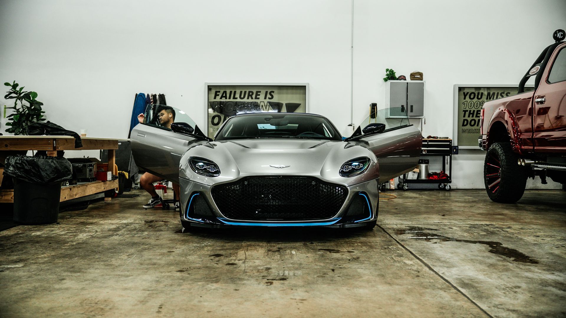 Silver sports car in a garage with open doors, a person in the car, and a red truck.
