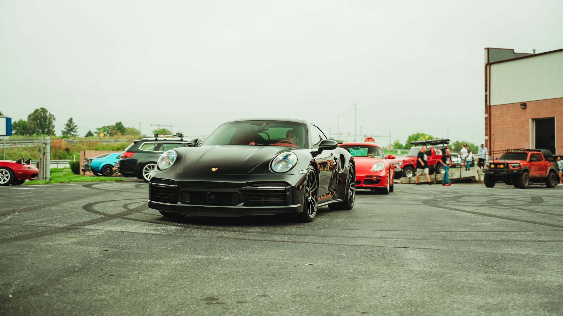 Black Porsche 911 at a car meet, surrounded by other vehicles. Gray pavement. Overcast sky.