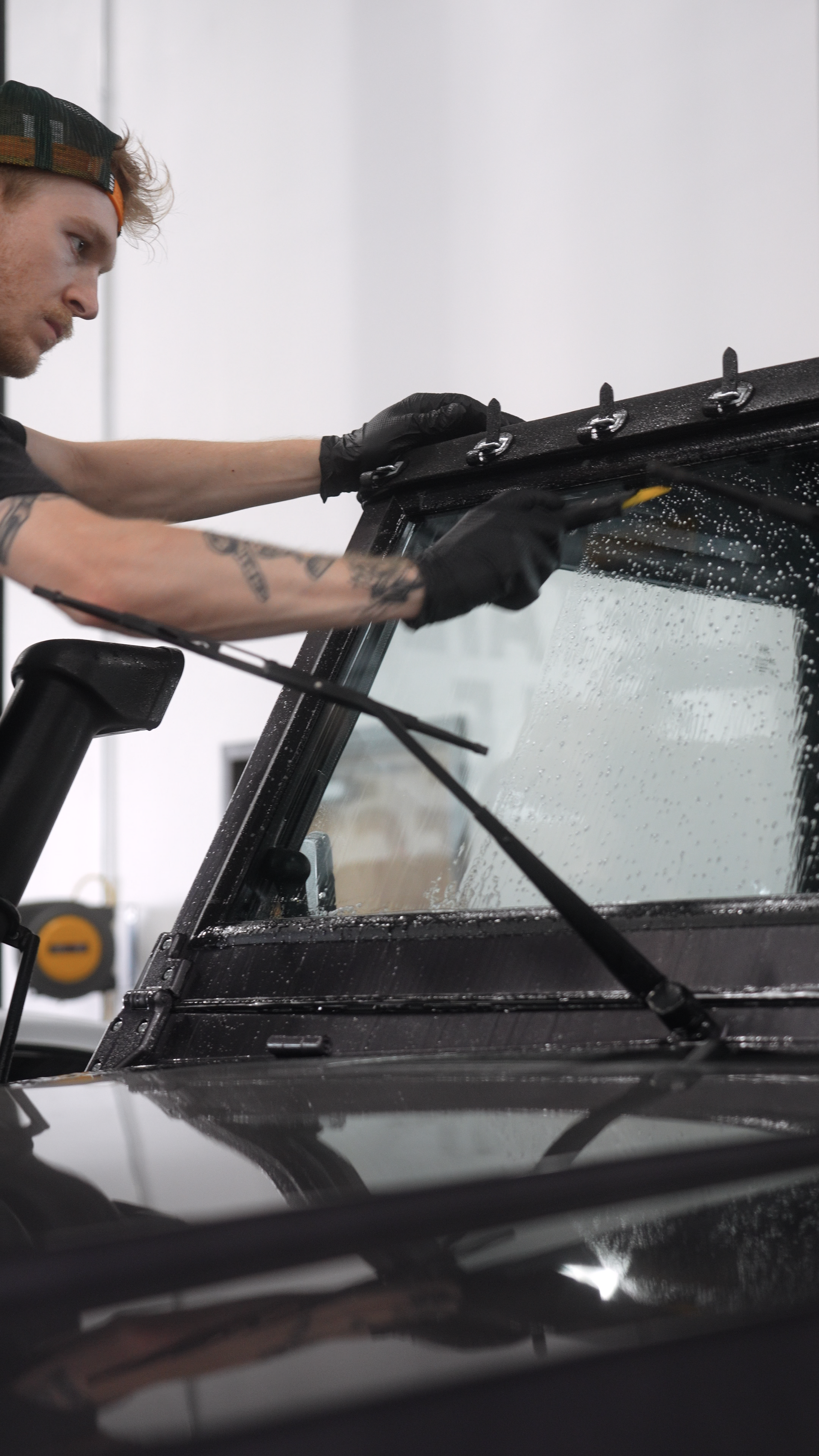 Person applying tint to a car windshield. Black gloves, cap, and vehicle. Interior setting.
