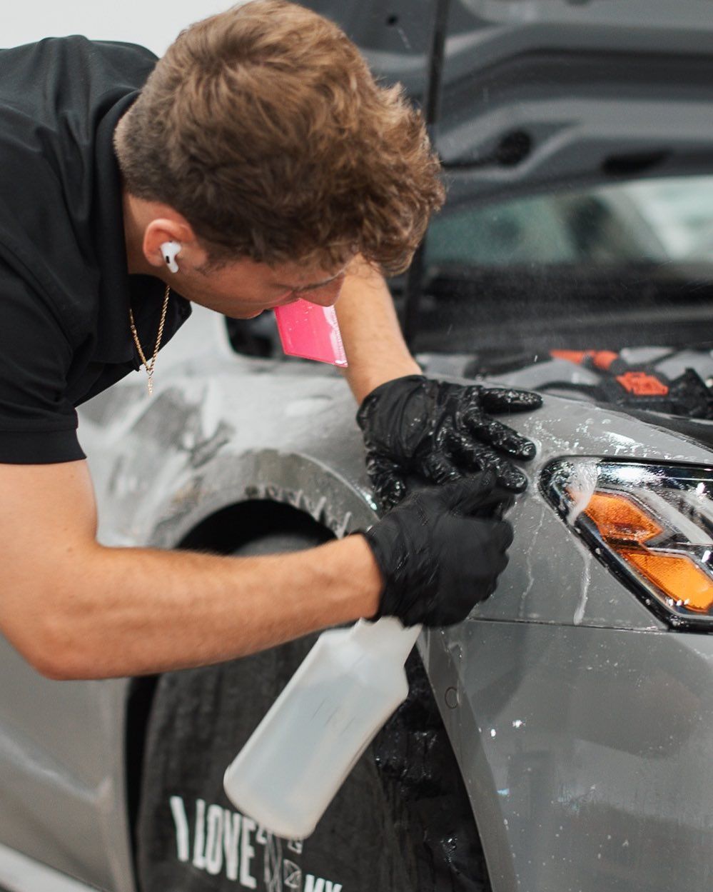 Person sprays liquid on a car's fender while wearing gloves; in a garage.
