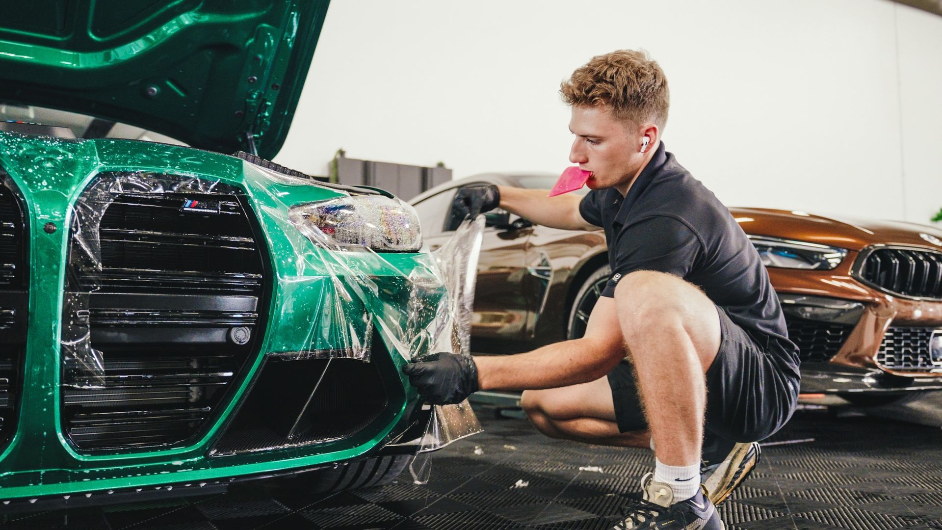 A man is kneeling down to apply protective film to the front of a green car.