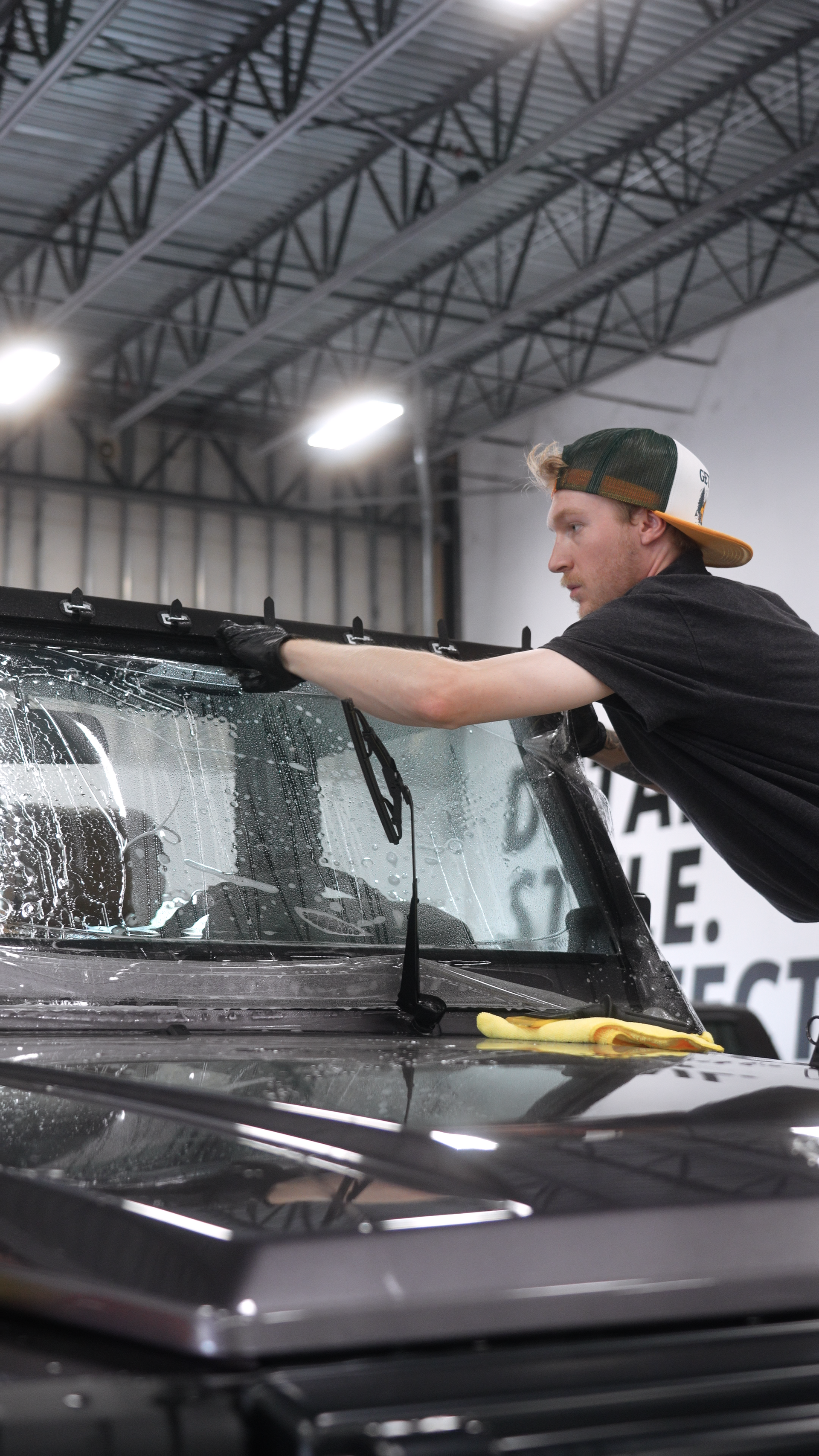 Man washing a black car's windshield with soapy water in a garage setting.