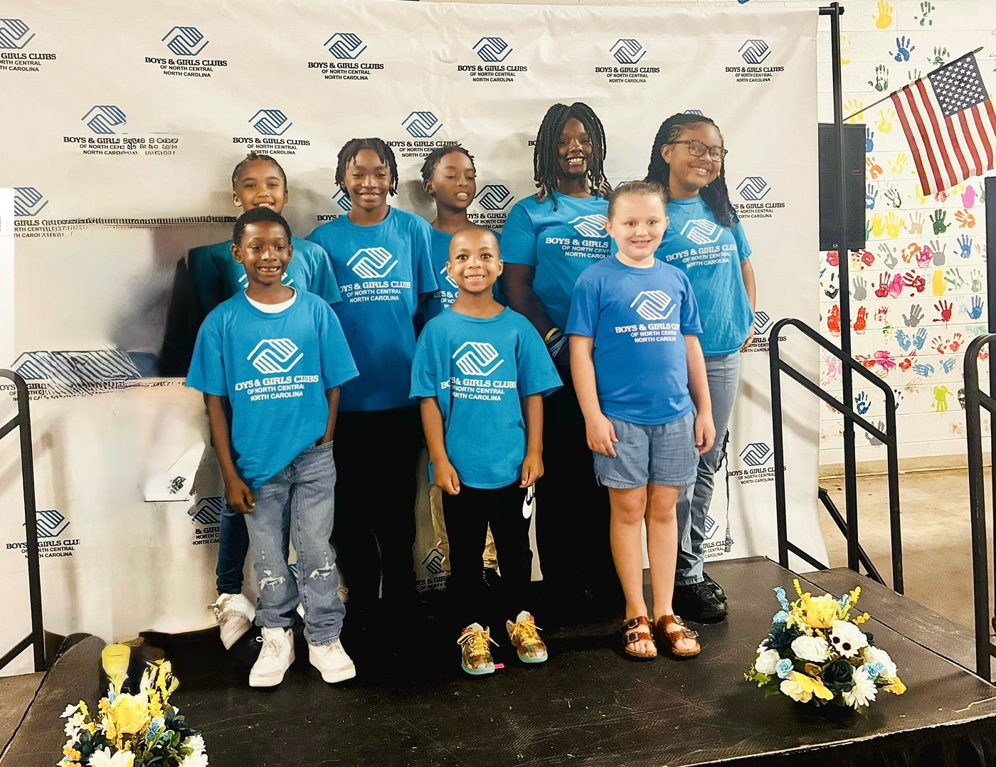 Six children in blue shirts pose on a small stage with a logo backdrop and floral arrangements.