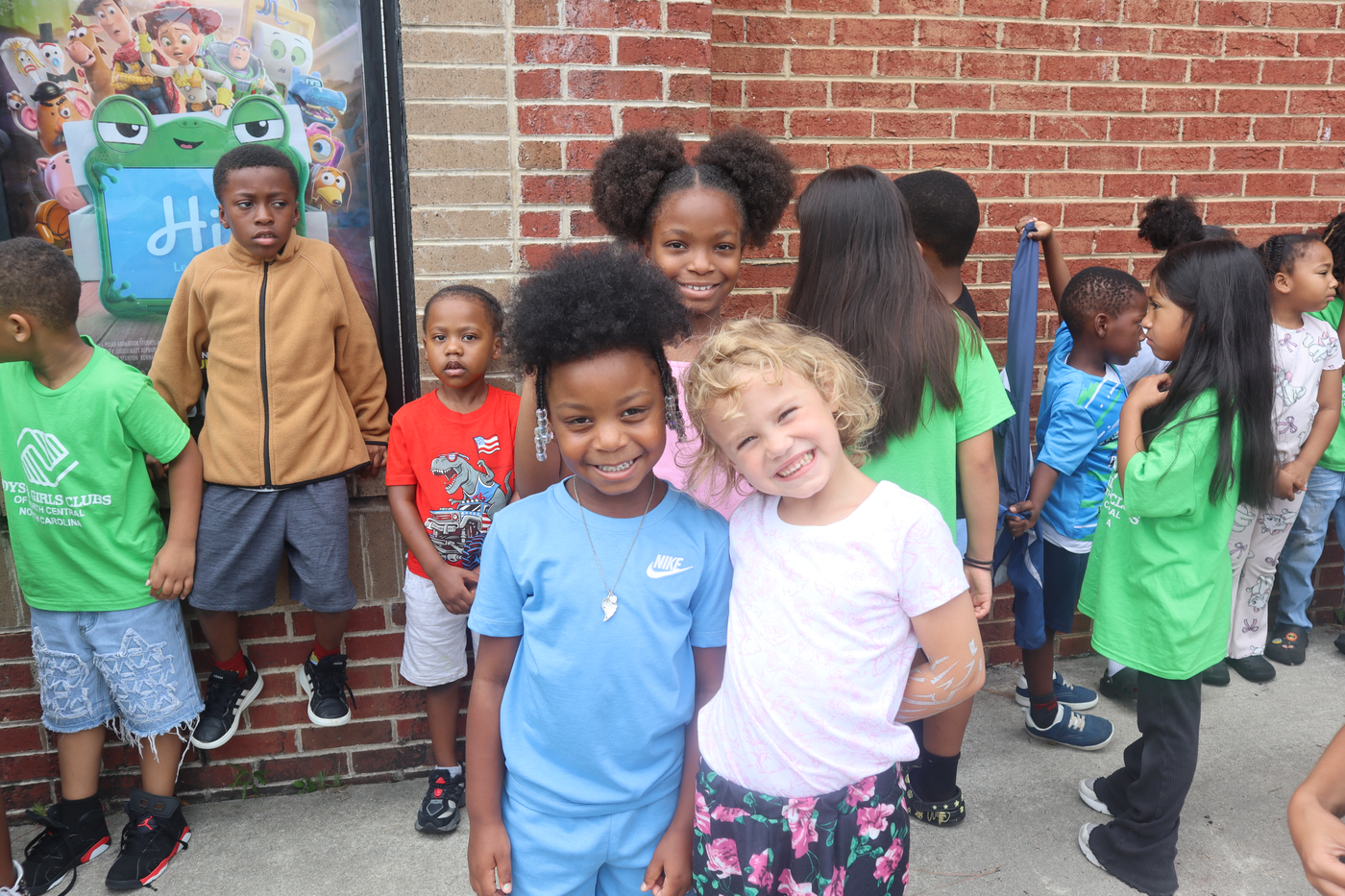 Children smiling and standing together outdoors in front of a brick wall and colorful mural