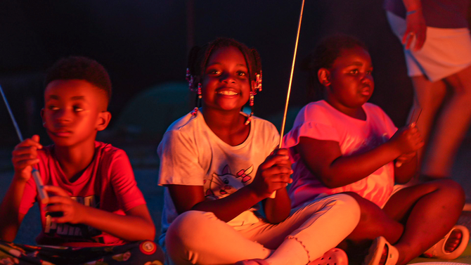Three children sitting on the ground by a warm orange campfire, smiling and clapping in the dark