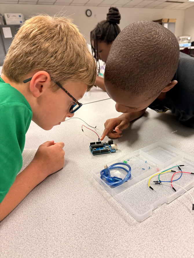 Two children lean over a small circuit board, working together at a classroom table.