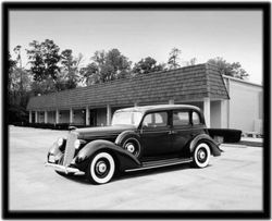 Black and white photo of a classic black car parked in front of a building with a shingled roof and white pillars.