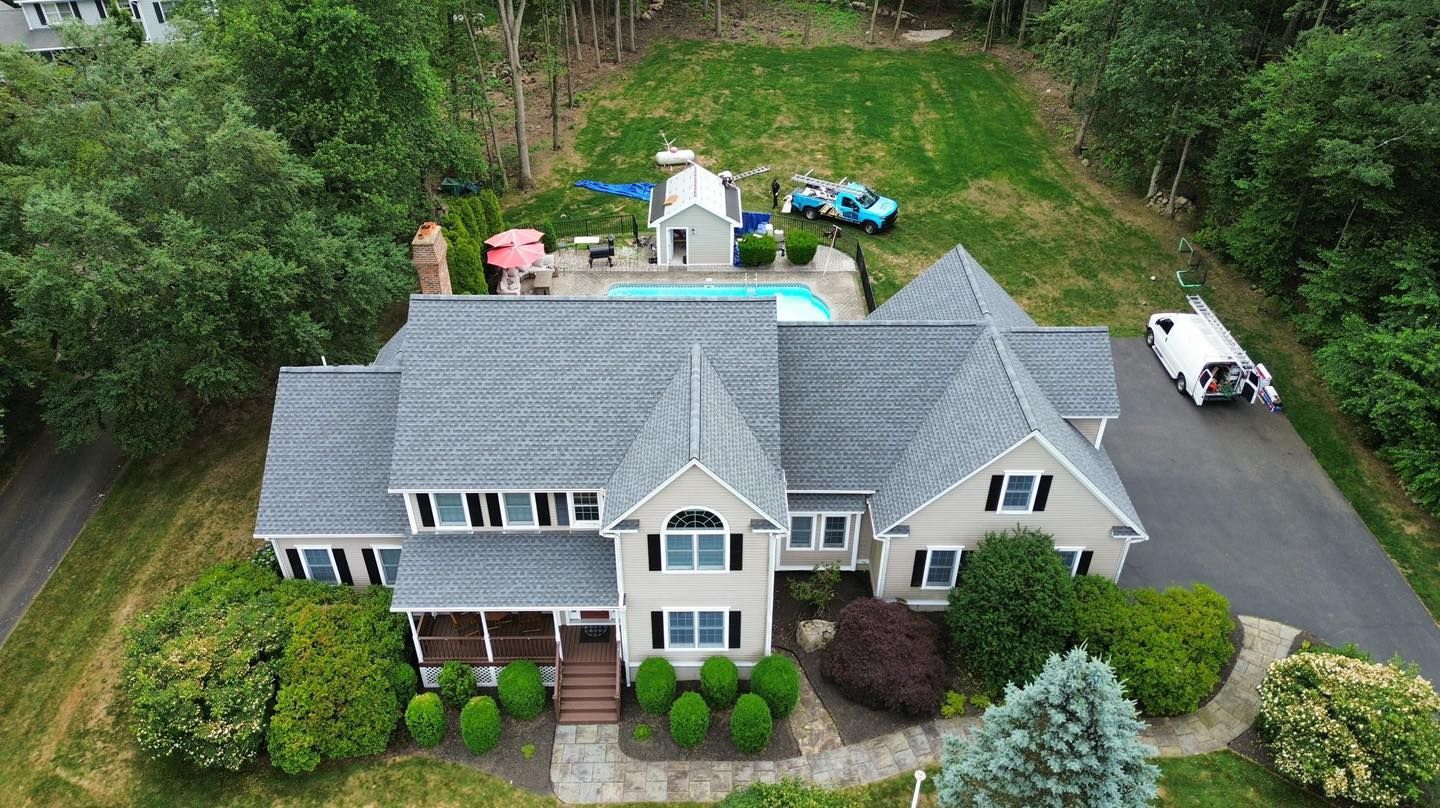 Aerial view of a gray suburban house with a driveway, front shrubs, and a wooded backyard.