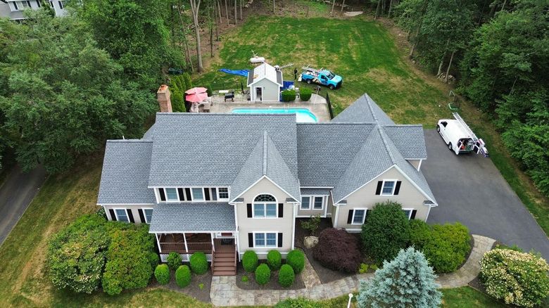 Aerial view of a gray suburban house with a driveway, front shrubs, and a wooded backyard.