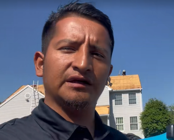 Man speaking in front of a house under construction; blue sky backdrop.