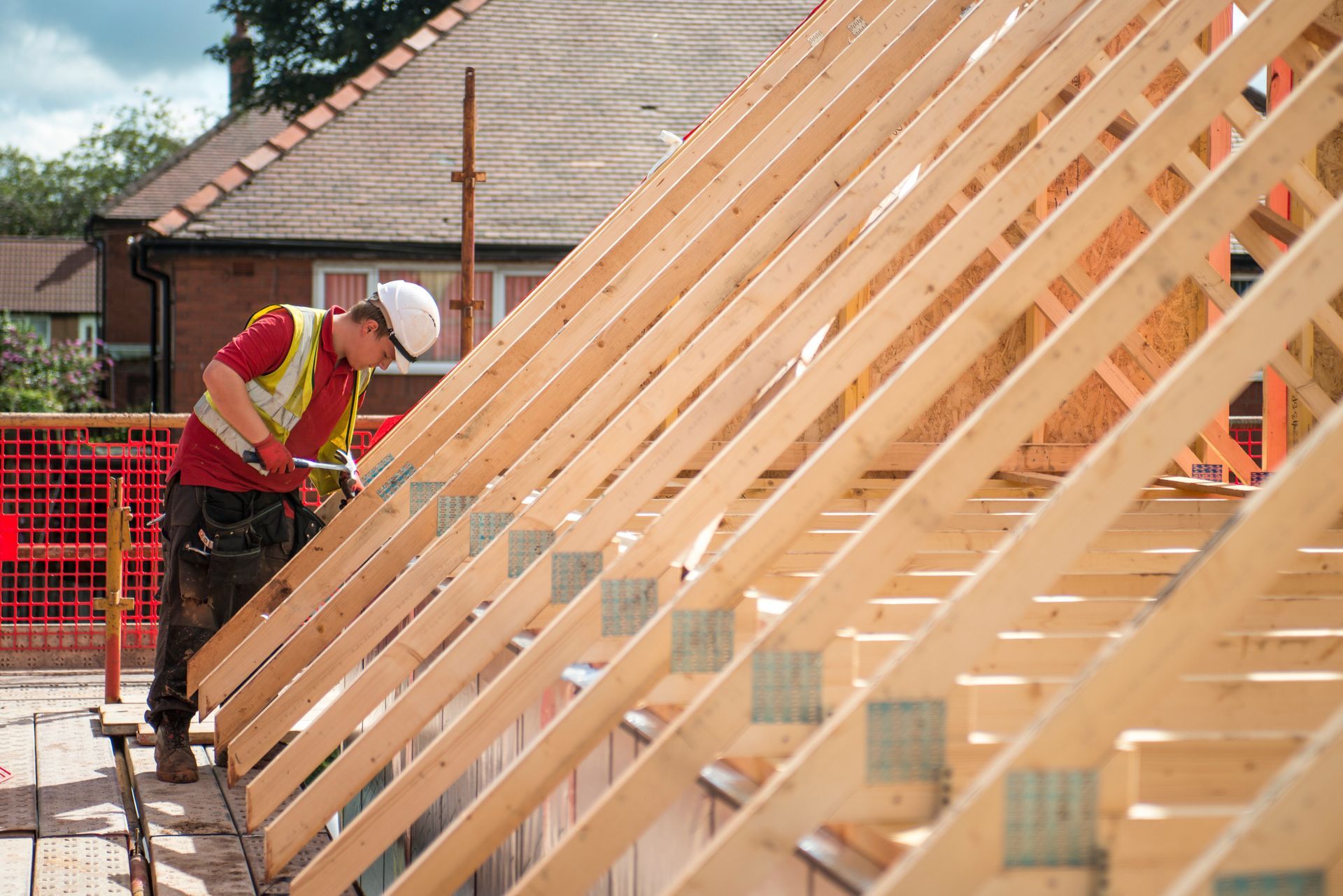 Construction worker in red vest and white hard hat framing a wooden house wall outdoors