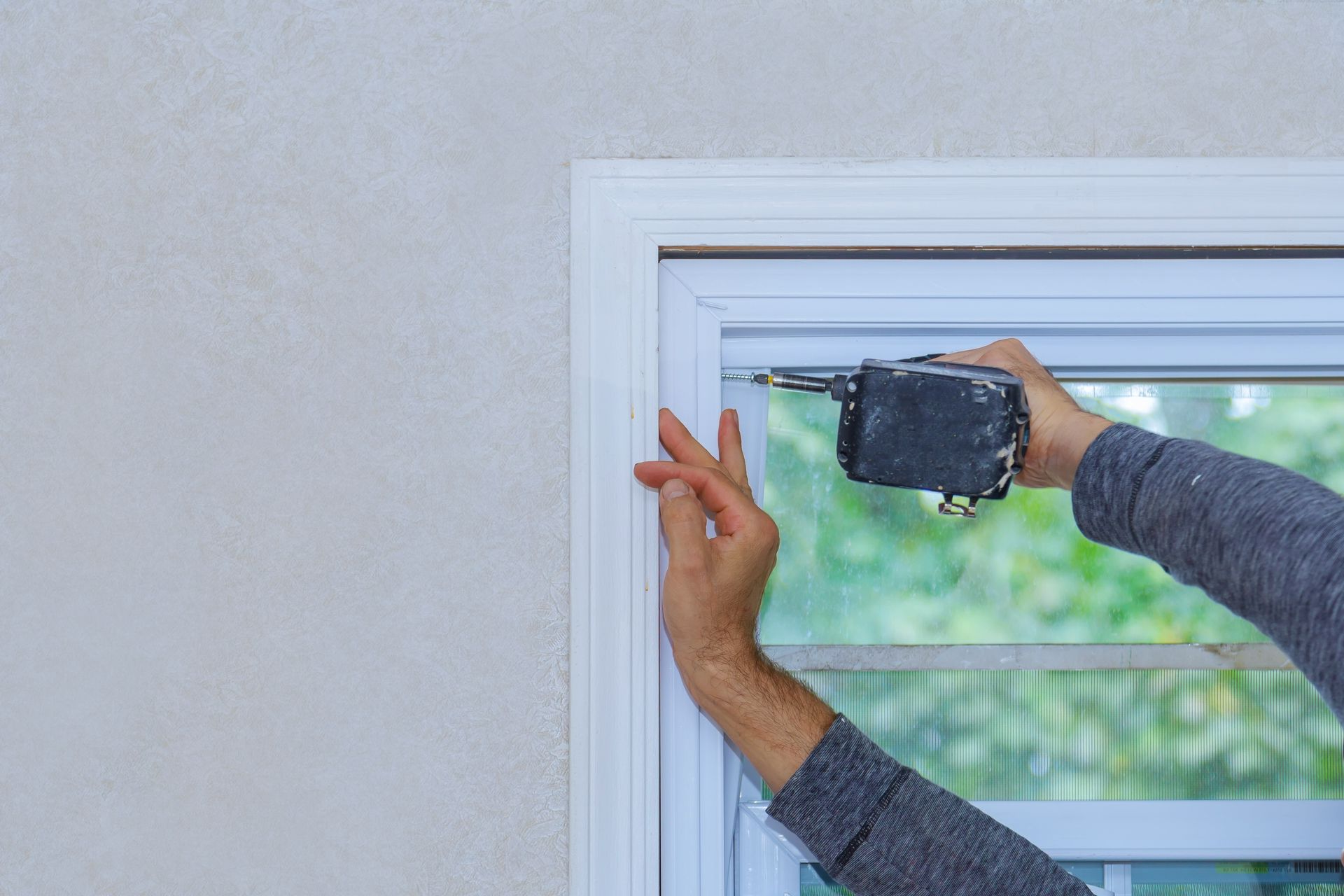 Person using a power drill to install a white window frame on a light gray wall.
