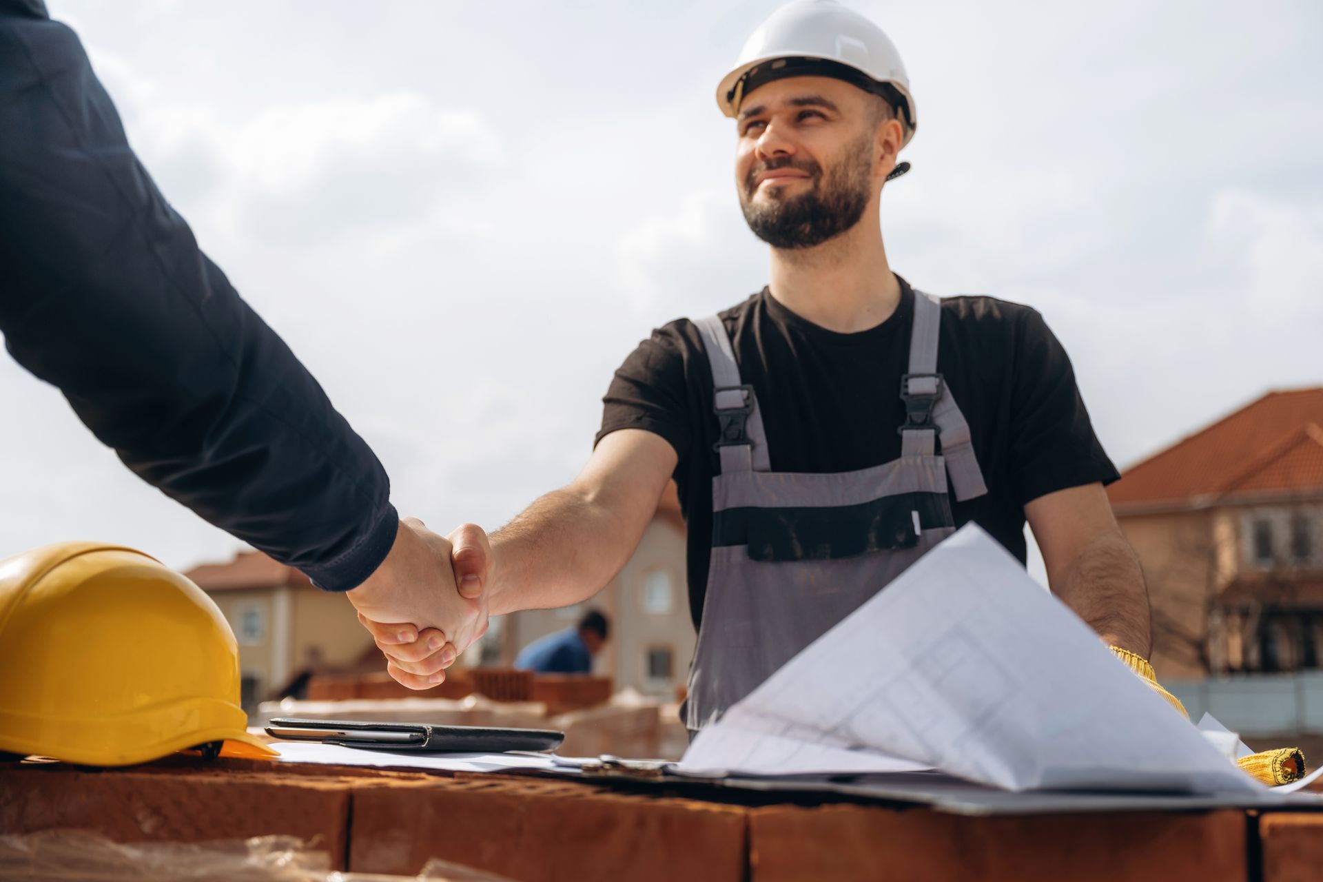 Construction worker shaking hands on a building site, with hard hats and blueprints on a table.