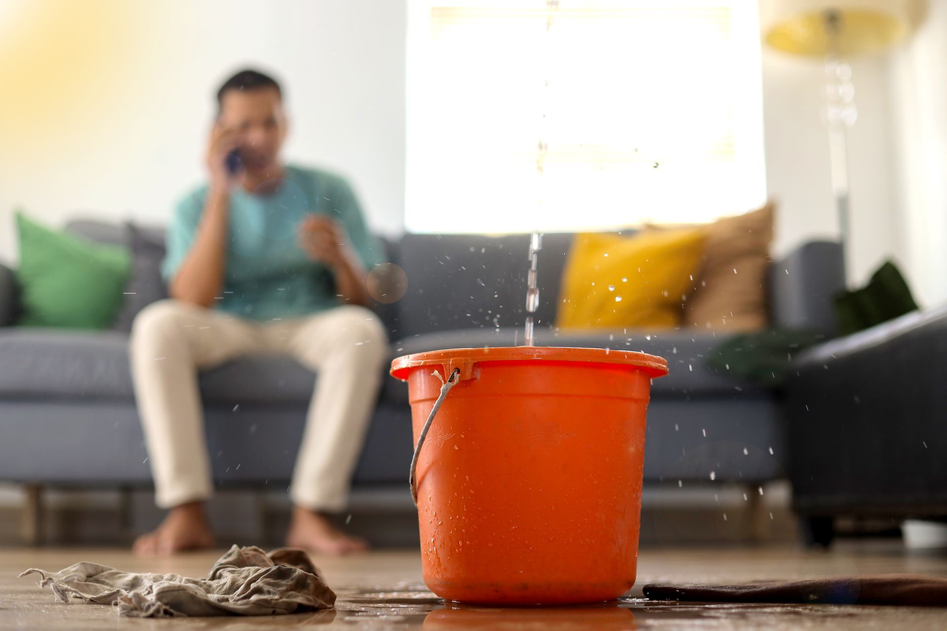 Orange bucket catching a roof leak in a living room, with a blurred person sitting on a couch behind it