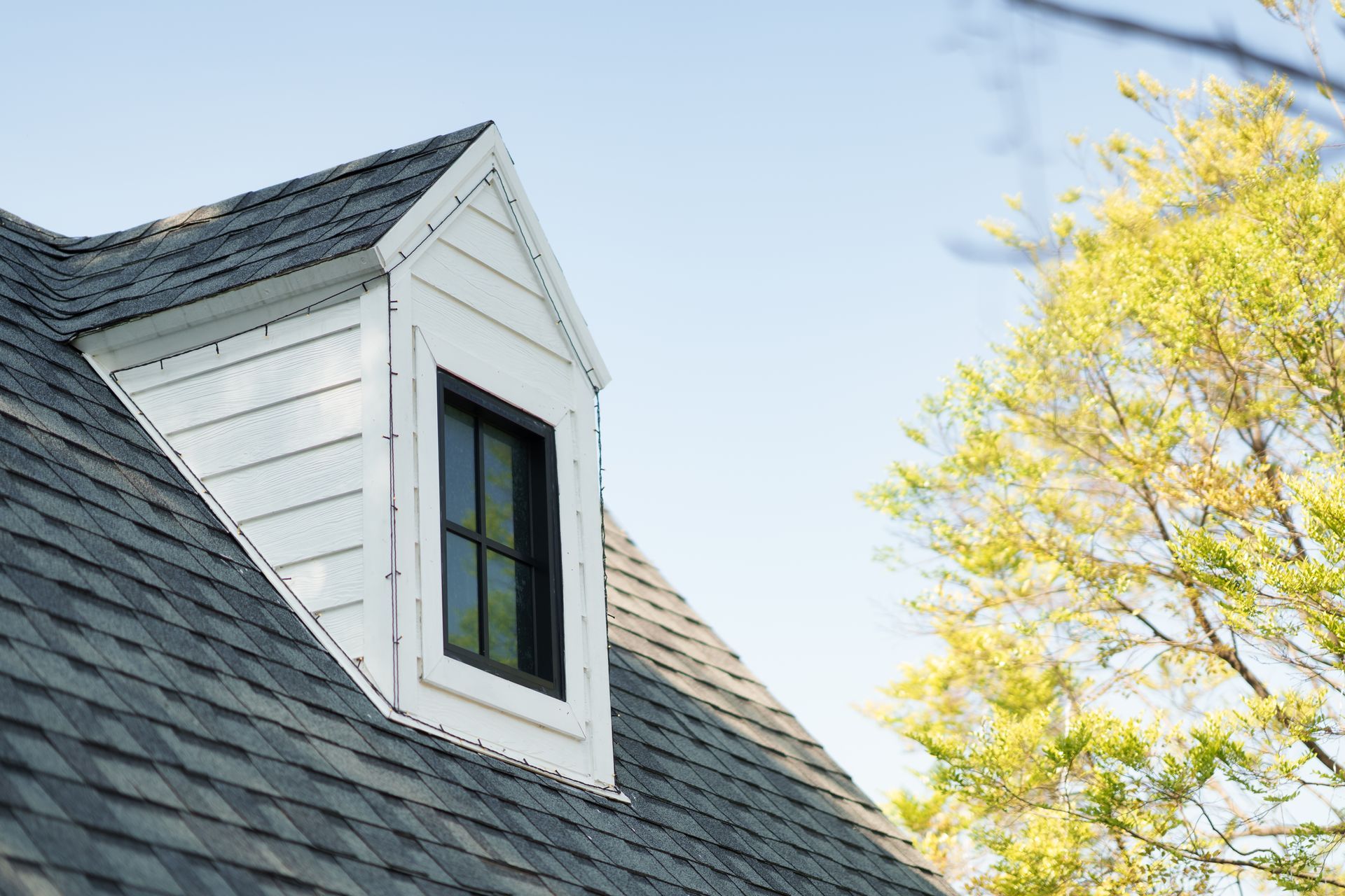 White dormer window on a gray shingled roof beside a yellow-leaved tree under a blue sky