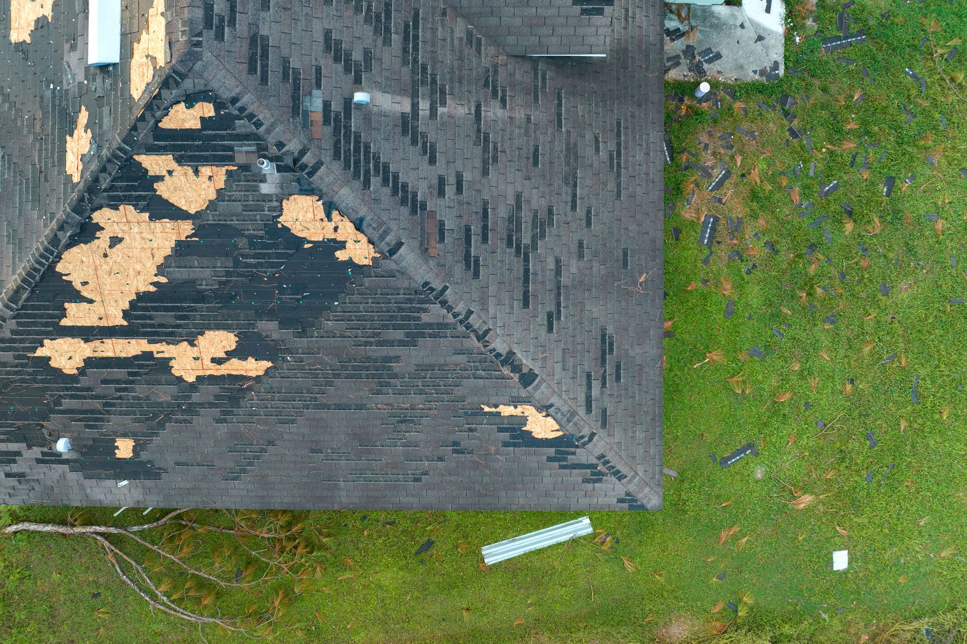 Aerial view of a roof with missing shingles beside a green grassy yard with scattered debris.
