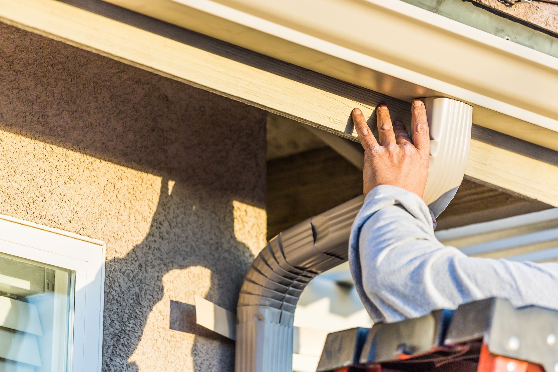 Person installing a downspout and gutter on a beige house exterior.