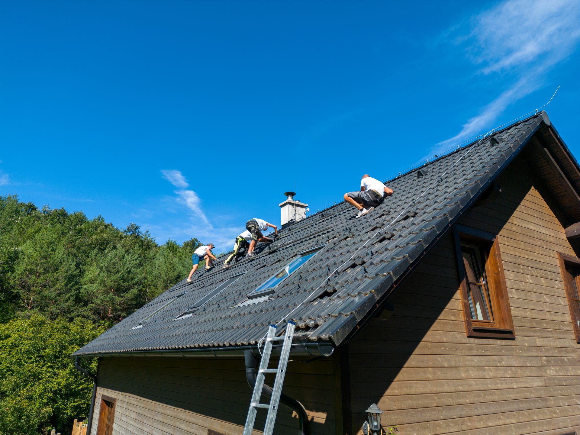 Workers repairing a dark roof on a wooden house under a bright blue sky