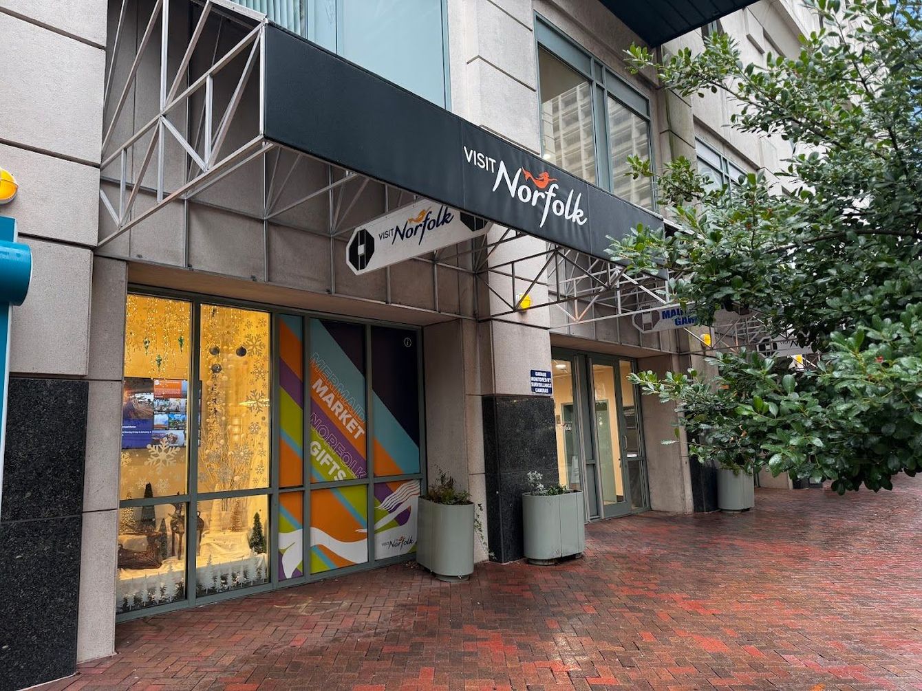 Entrance to The Norfolk building with a black awning, glass storefront, and potted plants on a brick sidewalk.