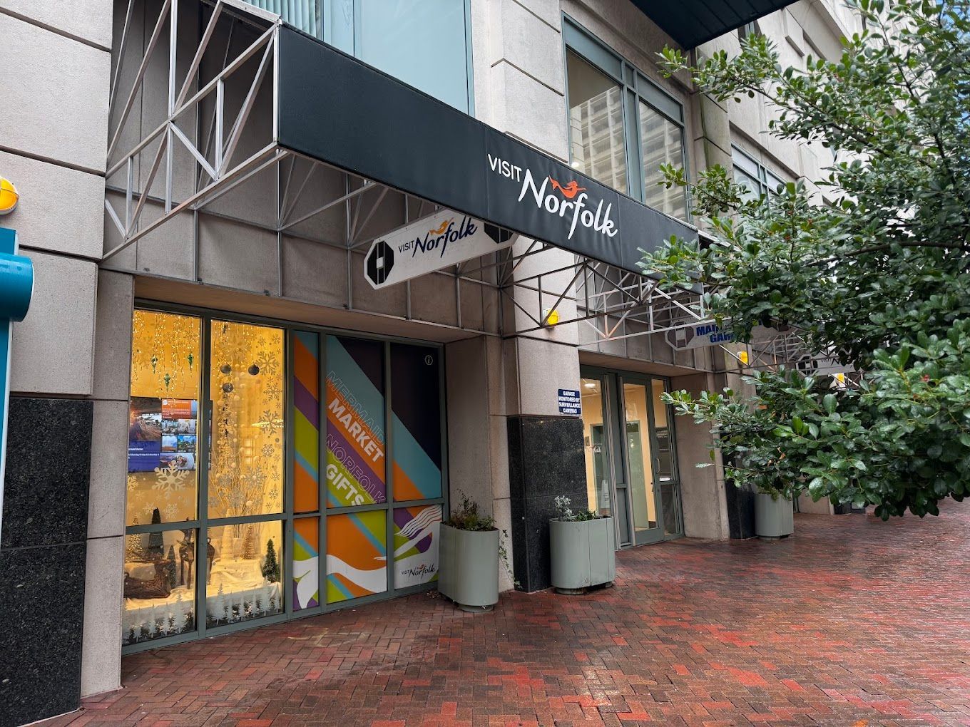 A street-level view of The Norfolk storefront entrance with floor-to-ceiling windows and large potted plants.
