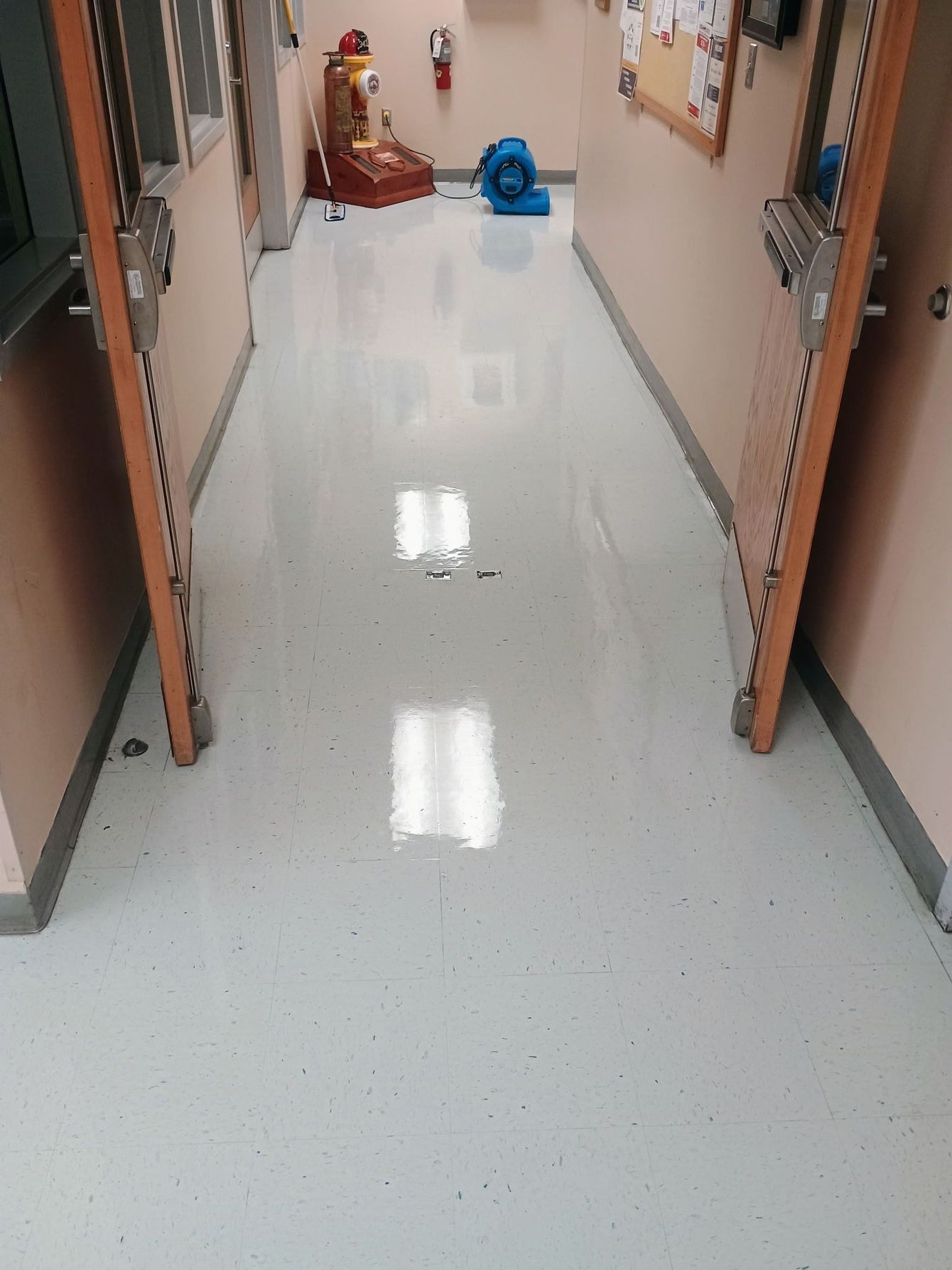 A polished hallway floor reflects ceiling lights, with a fire extinguisher and a blue air mover visible at the end.