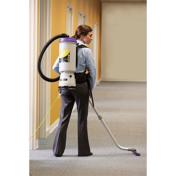 A person wearing a backpack vacuum cleaner walks down a carpeted hallway while cleaning the floor.