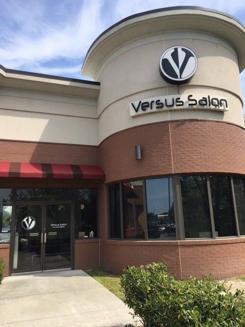 Exterior of Versus Salon, a building with a red-brick facade, a rounded wall, and a circular logo sign above the entrance.