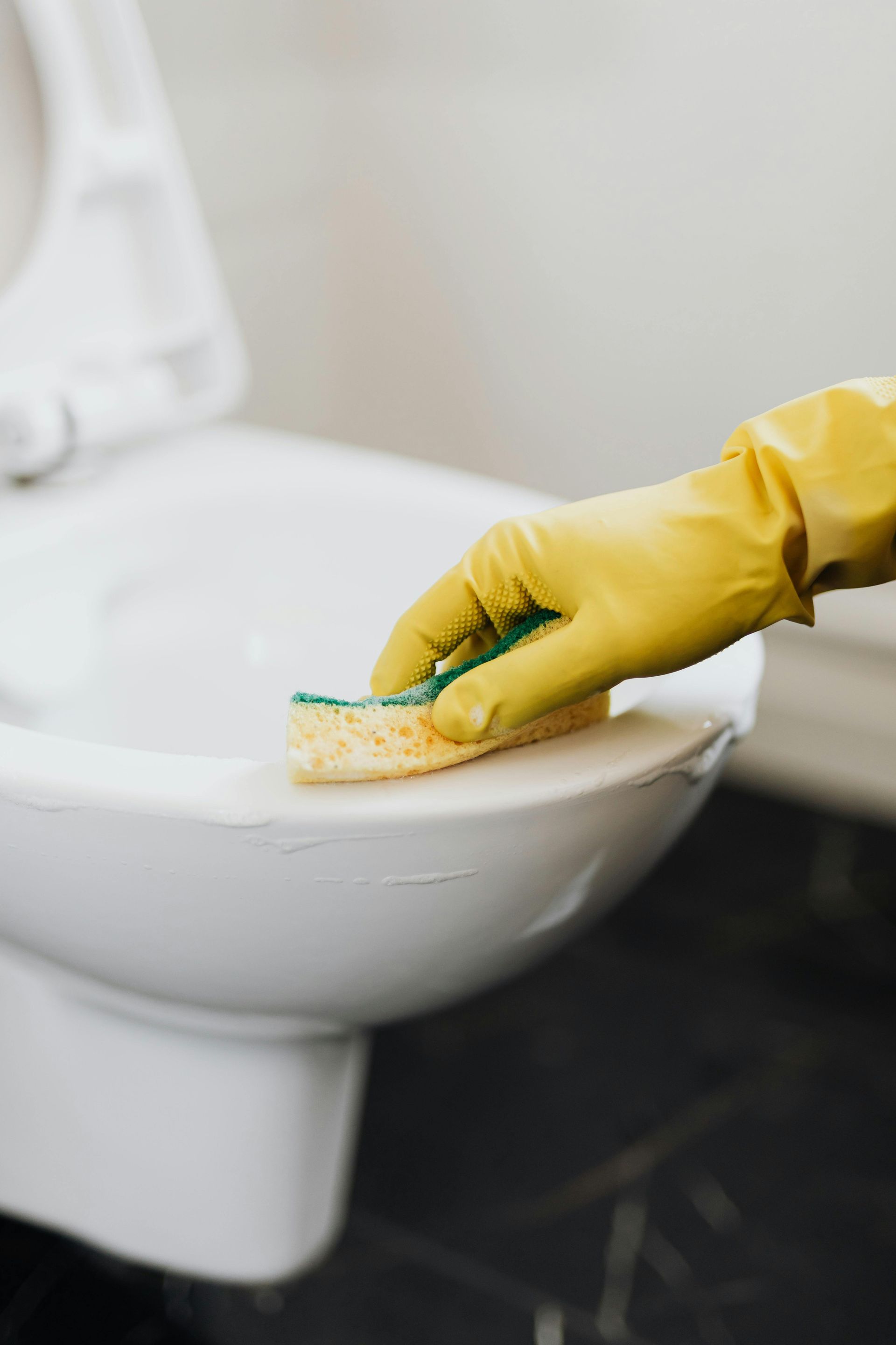 A hand wearing a yellow rubber glove uses a sponge to scrub the rim of a white porcelain toilet.