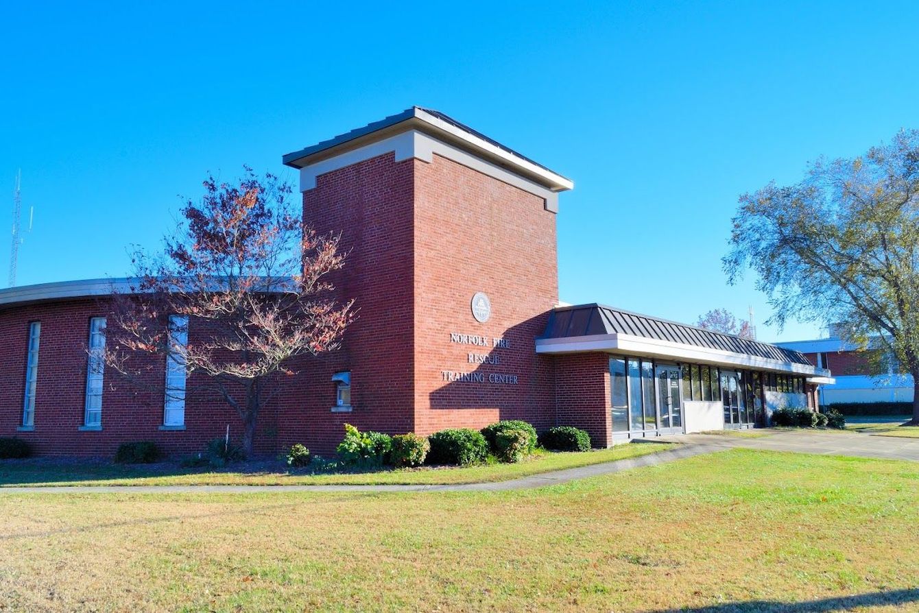 A red brick building with a central tower and flat roof, set against a bright blue sky with trees and a grassy lawn.