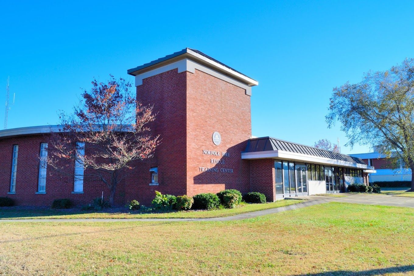 A red brick building with a central tower and flat roof, set against a bright blue sky with trees and a grassy lawn.